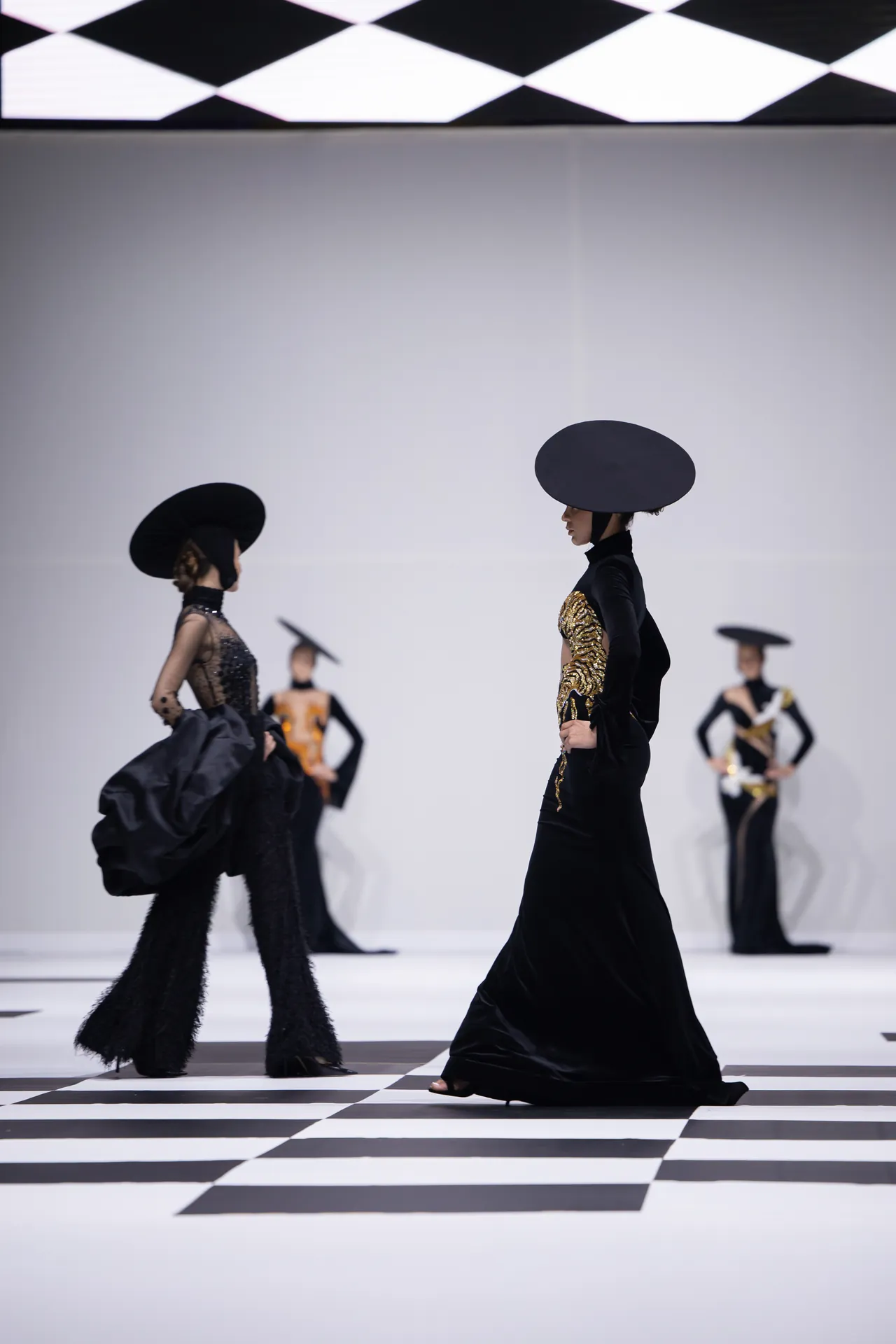 Models walk a fashion runway in black gowns with oversized circular hats on a bold black-and-white checkered floor. A minimalist white backdrop emphasizes the dramatic silhouettes.
