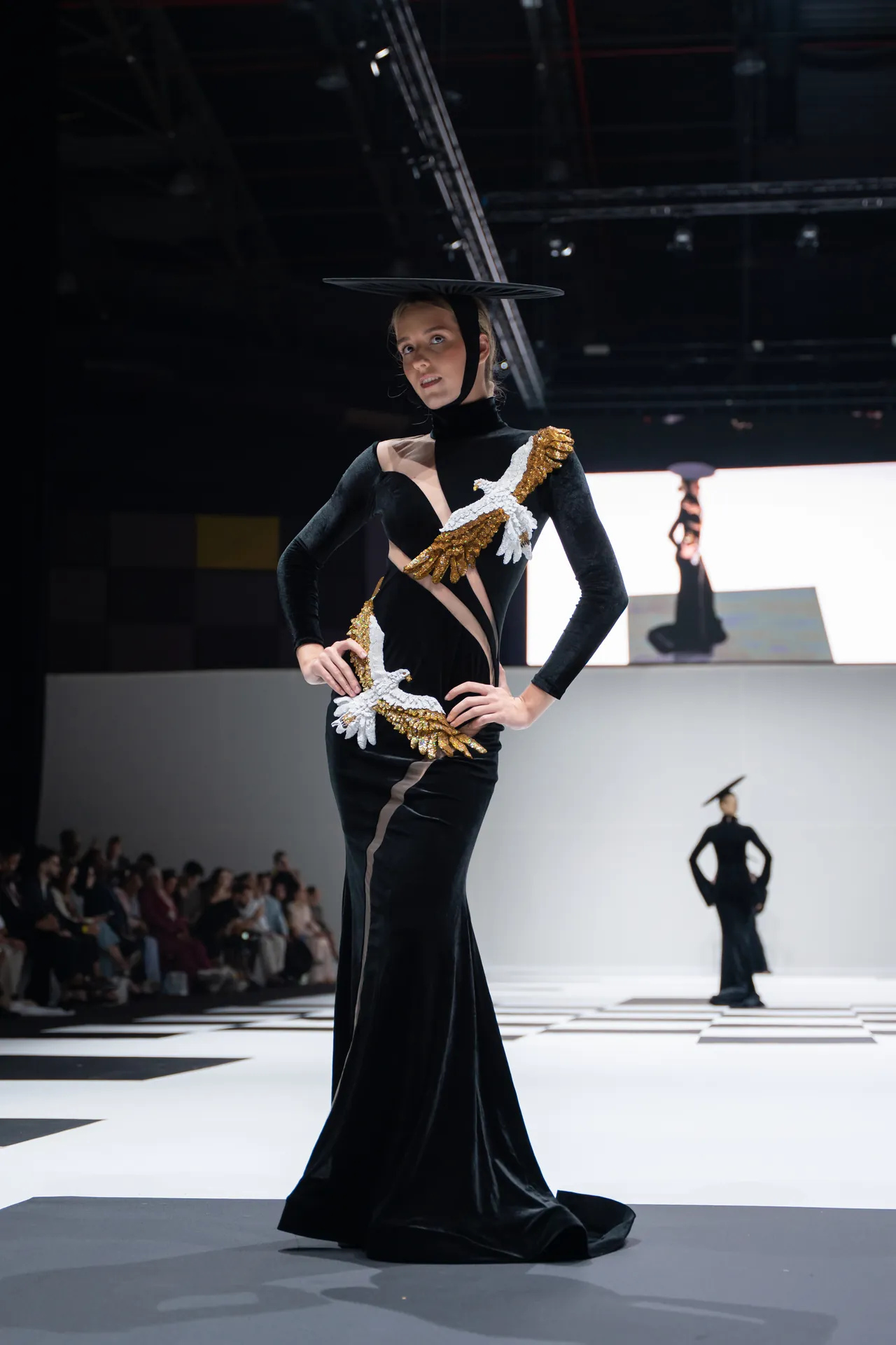 Runway model in a black velvet dress with gold-and-white feather embellishment, balancing a large circular hat on her head.