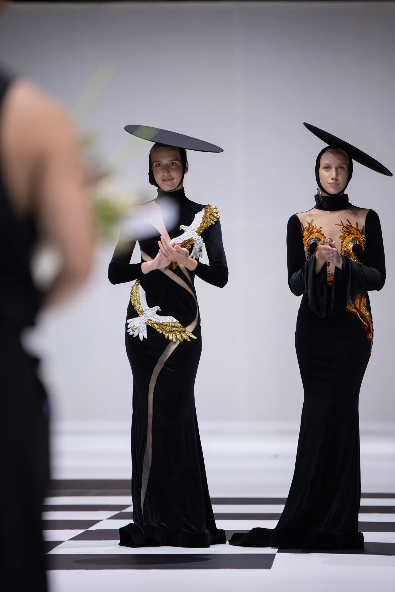 Two models in black gowns with gold embroidery stand on a black-and-white checkerboard floor, wearing oversized circular hats on their heads? Note: fix phrasing - Two models in black gowns with gold embroidery stand on a black-and-white checkerboard floor, wearing oversized circular hats on their heads.