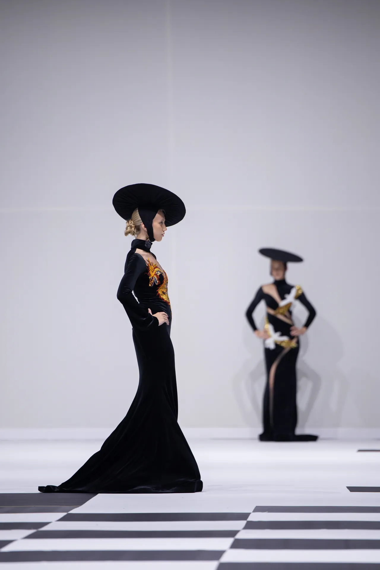 Model on a runway in a long black gown with a large circular hat, another model in the background wearing a similar outfit.