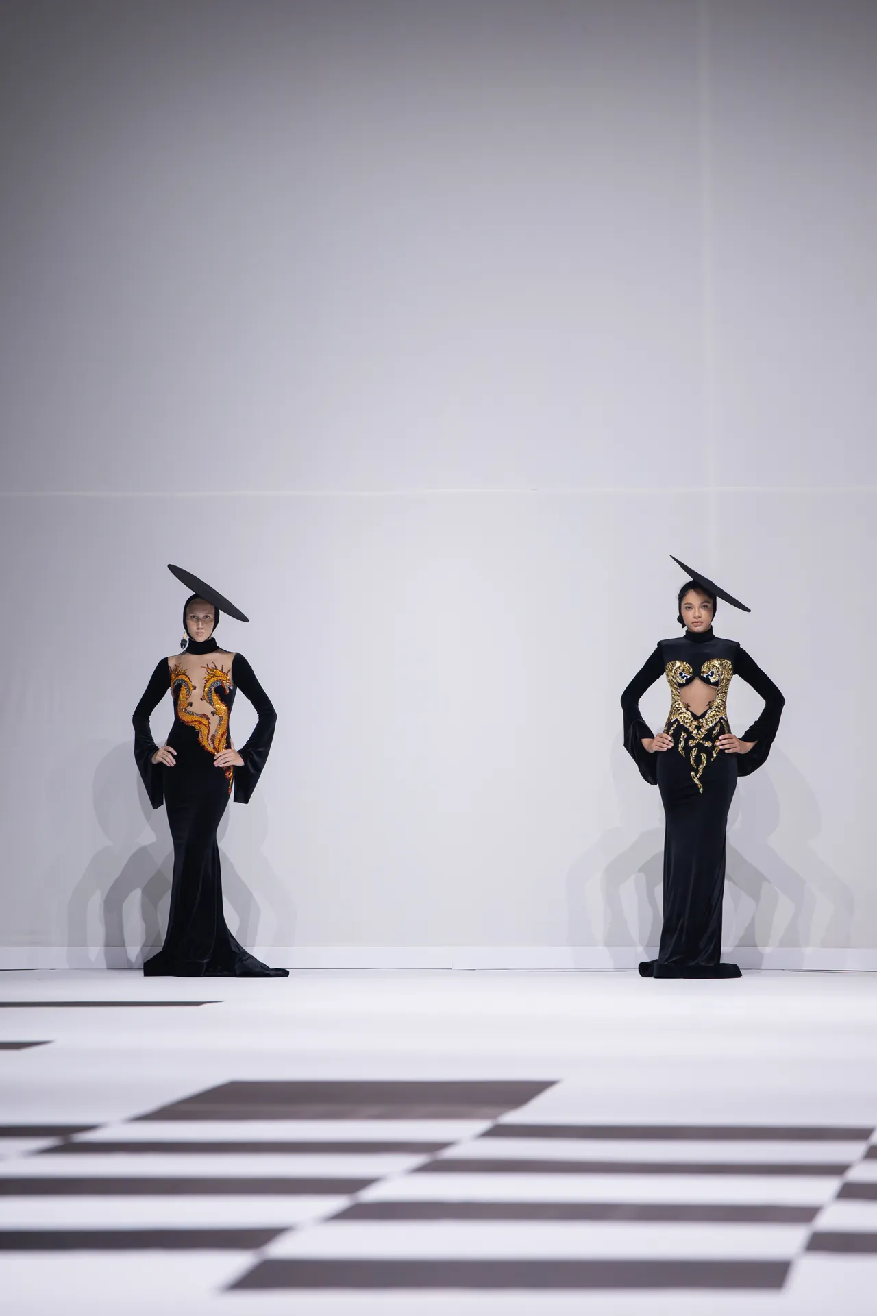 Two models in black, floor-length gowns with gold embroidery stand on a white runway, each wearing a wide, tilted black hat for a dramatic fashion show look.