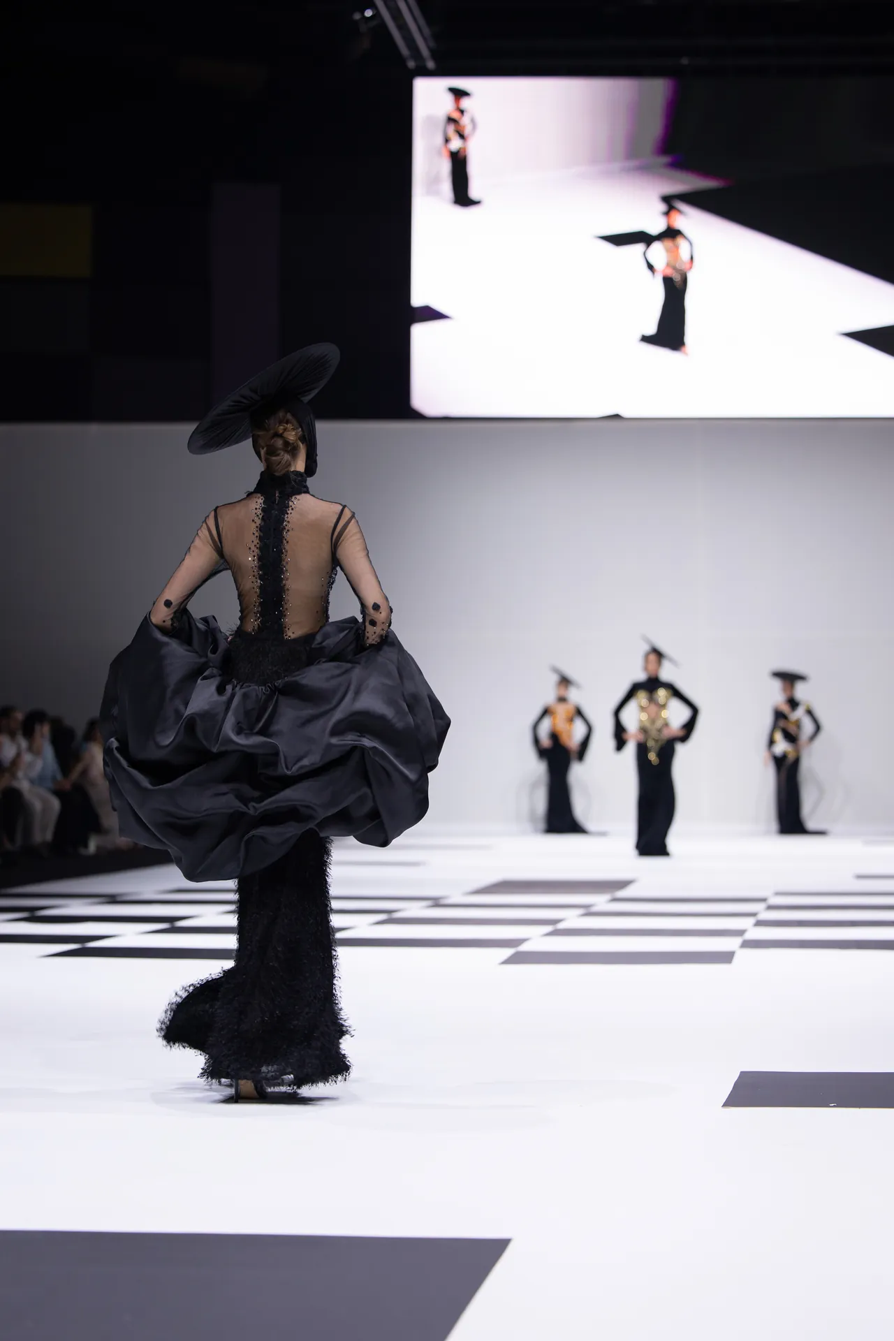 Back view of a model in a dramatic black gown with sheer panels and a wide hat, walking a white checkerboard runway at a fashion show.
