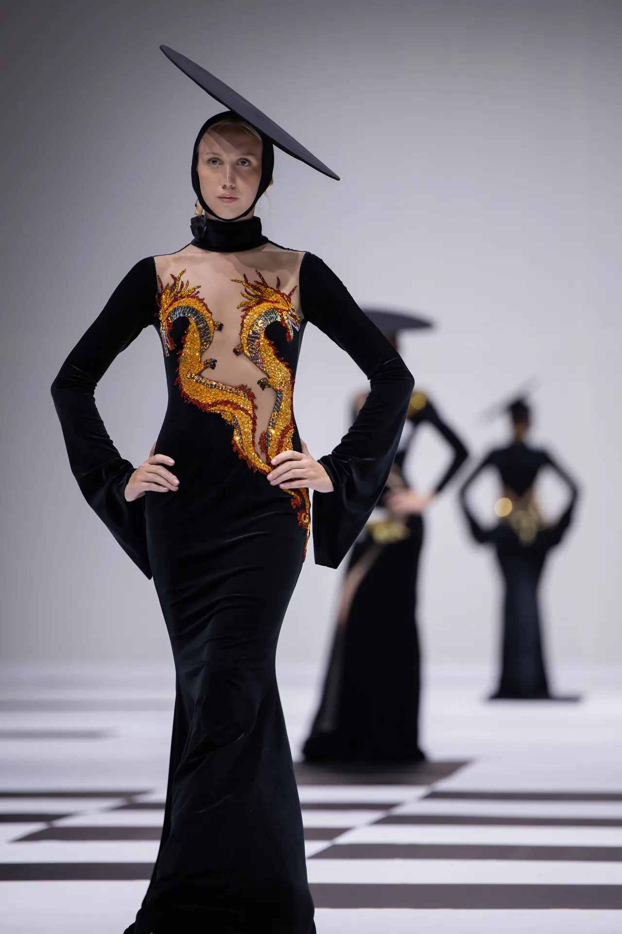 Model walking a runway in a black velvet gown with gold dragon embroidery and a large tilted black hat.