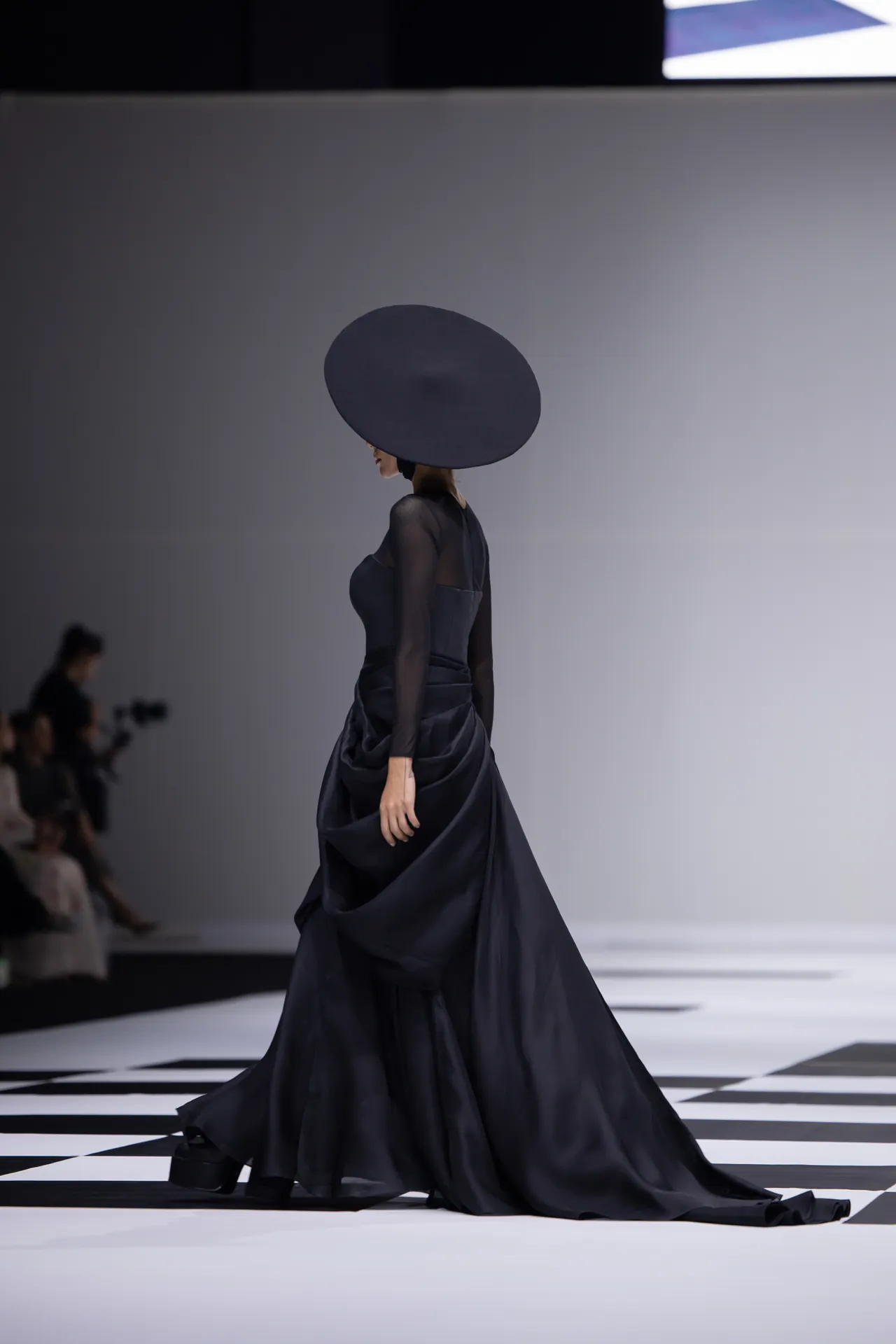 Model walking a runway in a dark, floor-length gown with a large circular brim hat, side profile view.