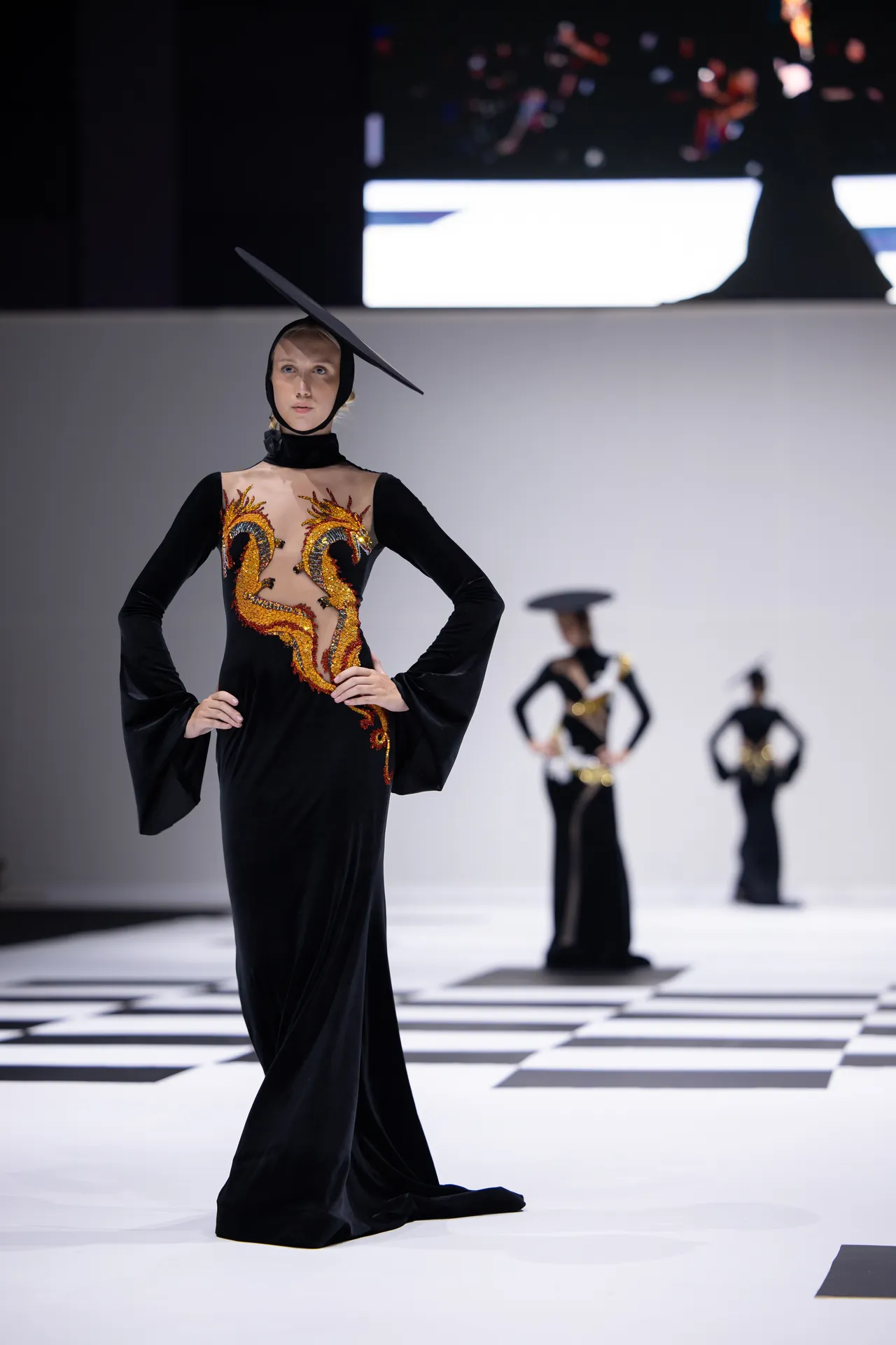 Model on a fashion runway wearing a black gown with sheer bodice and gold dragon embroidery, plus a sculptural tilted hat/ headpiece.