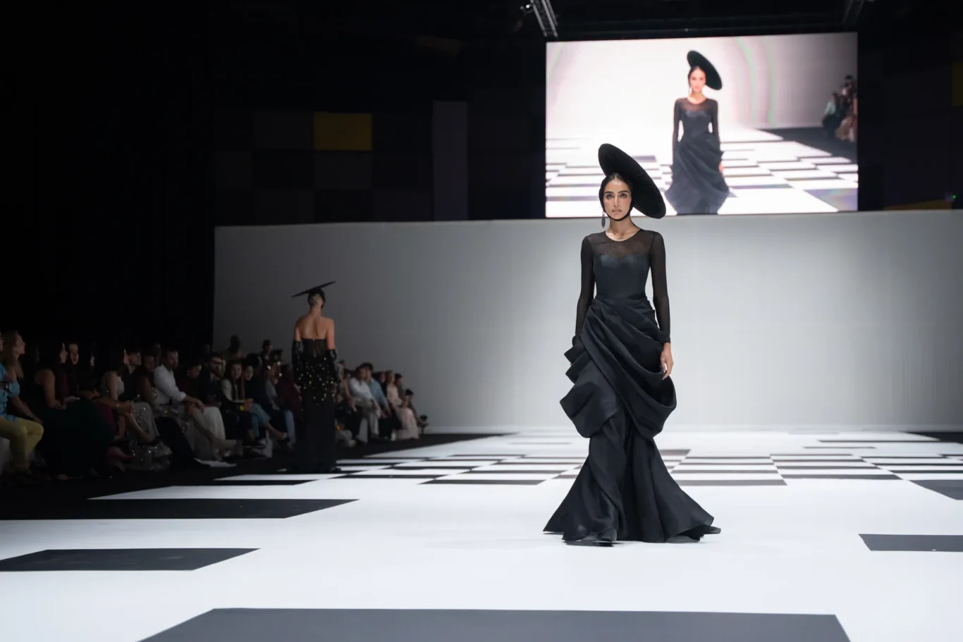 Model walks a white and black checkered runway in a black sheer gown with a large sculptural hat; audience watches from the left.