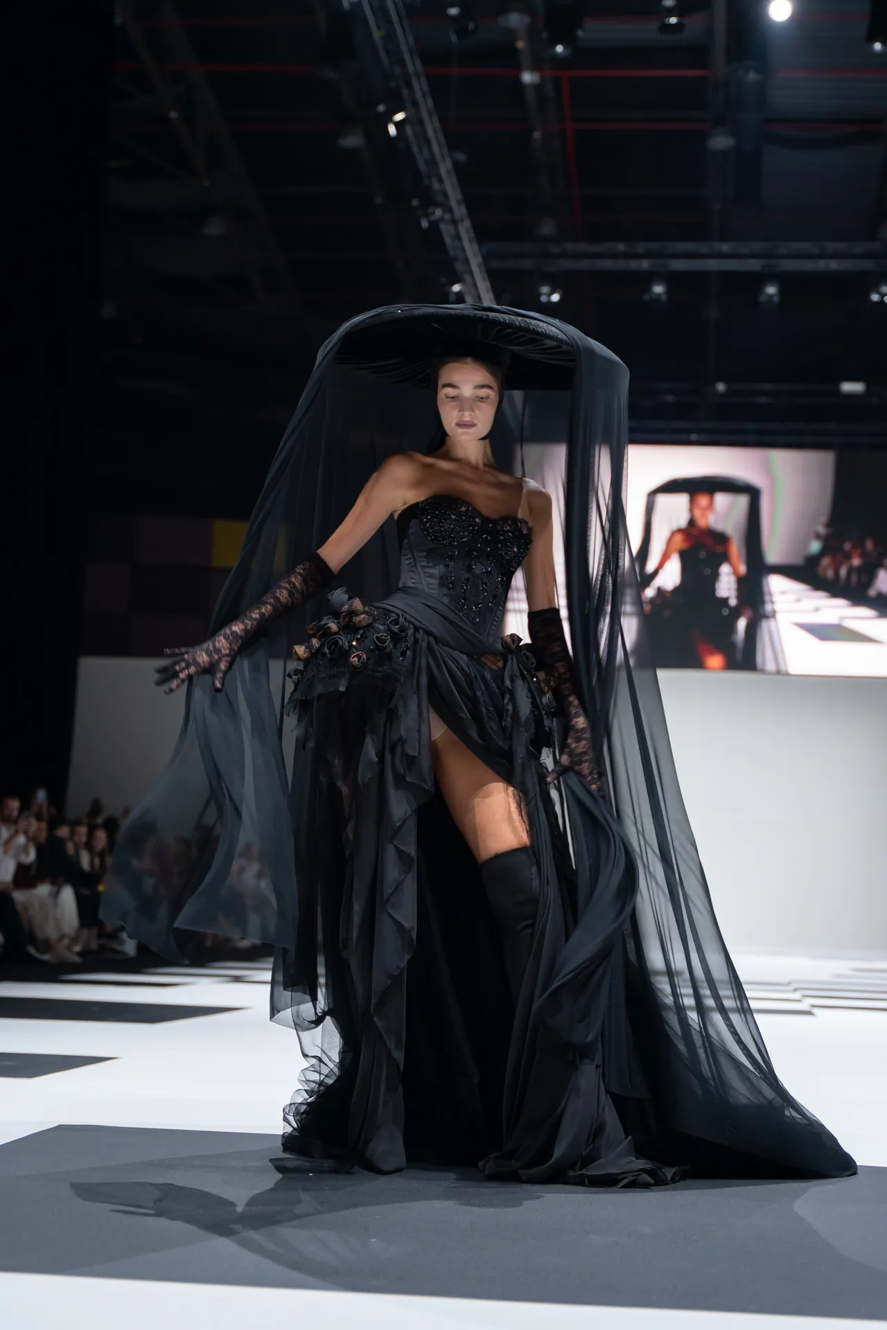 Model on a runway wearing a dramatic black gown with a long black veil cape and lace gloves.