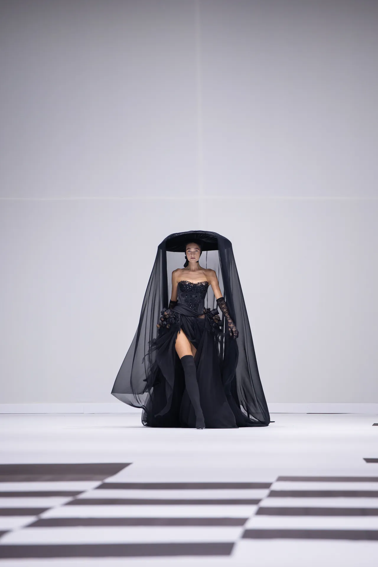 Model walking a runway in a black, strapless gown with a dramatic sheer cape and long gloves on a geometric black-and-white floor.