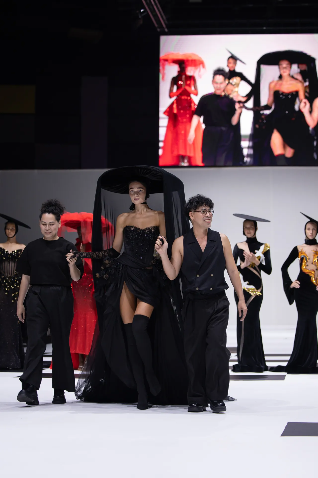 Models walk a fashion show runway in black outfits; central female in a sheer black gown with cape, a man in a black vest beside her; red dresses on screen backdrop.