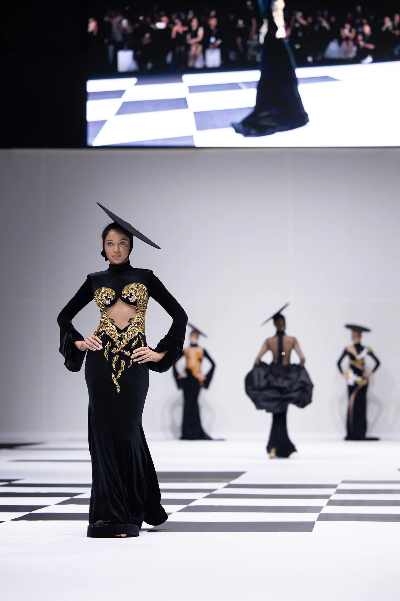 Model walks the runway in a black gown with gold embroidered detailing and a tilted large black hat; checkered floor and other models in background.