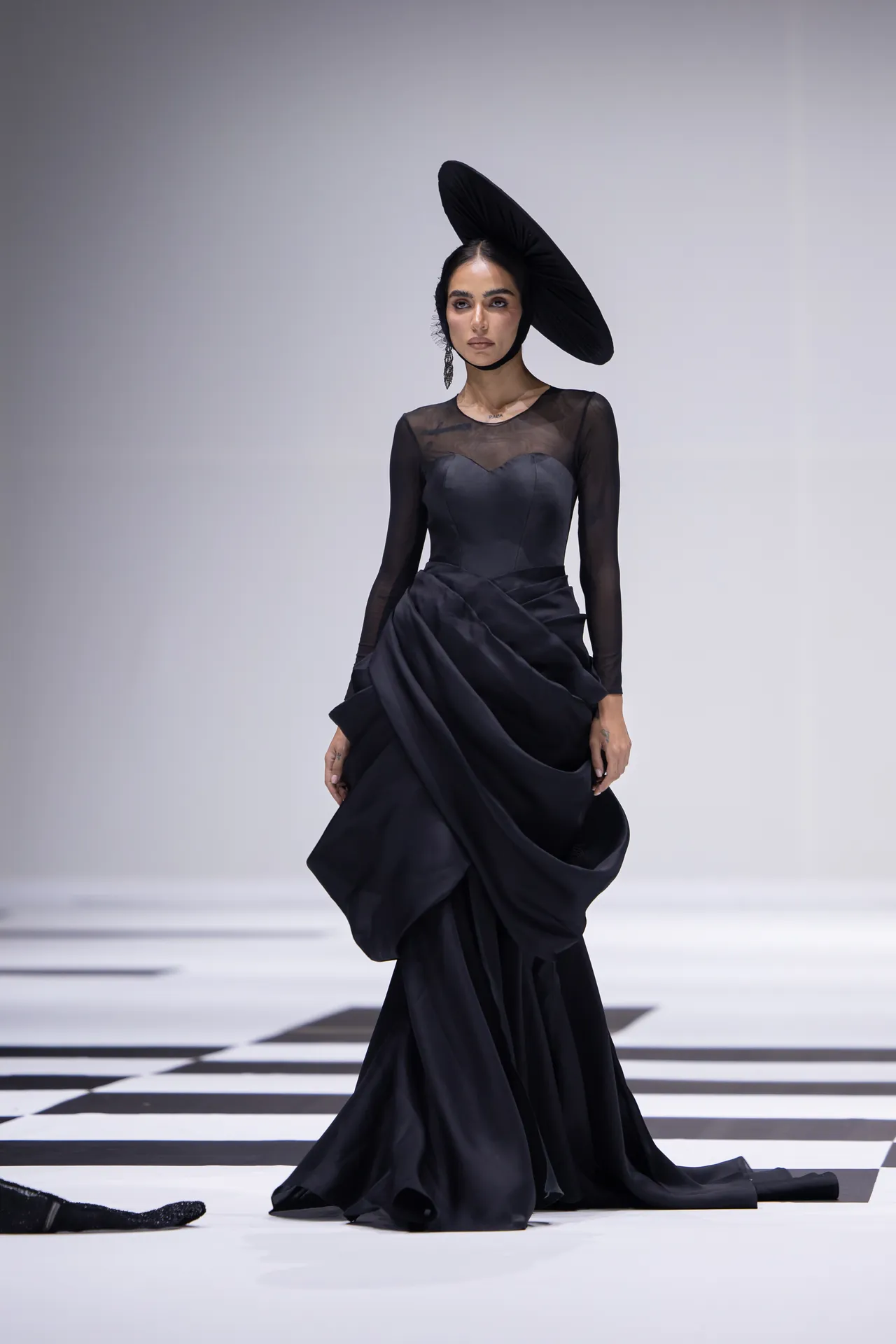 Model walking a fashion show in a dramatic black gown with sheer sleeves and a large circular black hat on a black-and-white checkered runway.