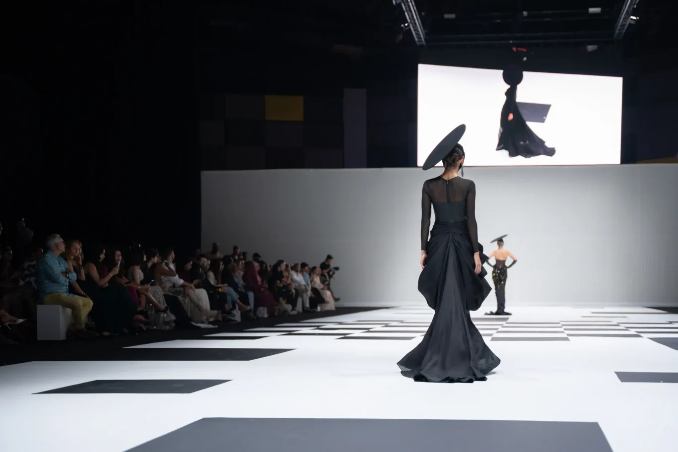 Model in a long black gown and oversized tilted hat walks a black-and-white checkered runway; audience seated along the left.