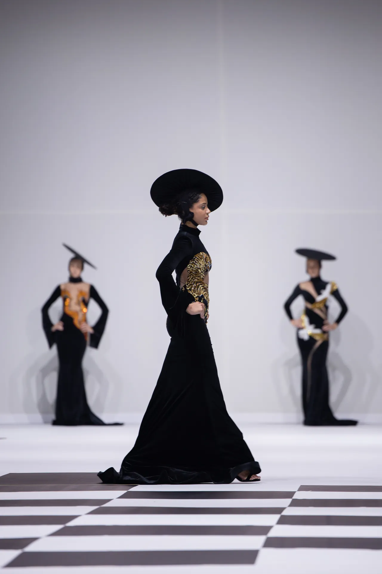 Female model walks the runway in a black velvet gown with gold embroidery and a wide black hat, on a black-and-white checkered floor, with other models in the blurred background.