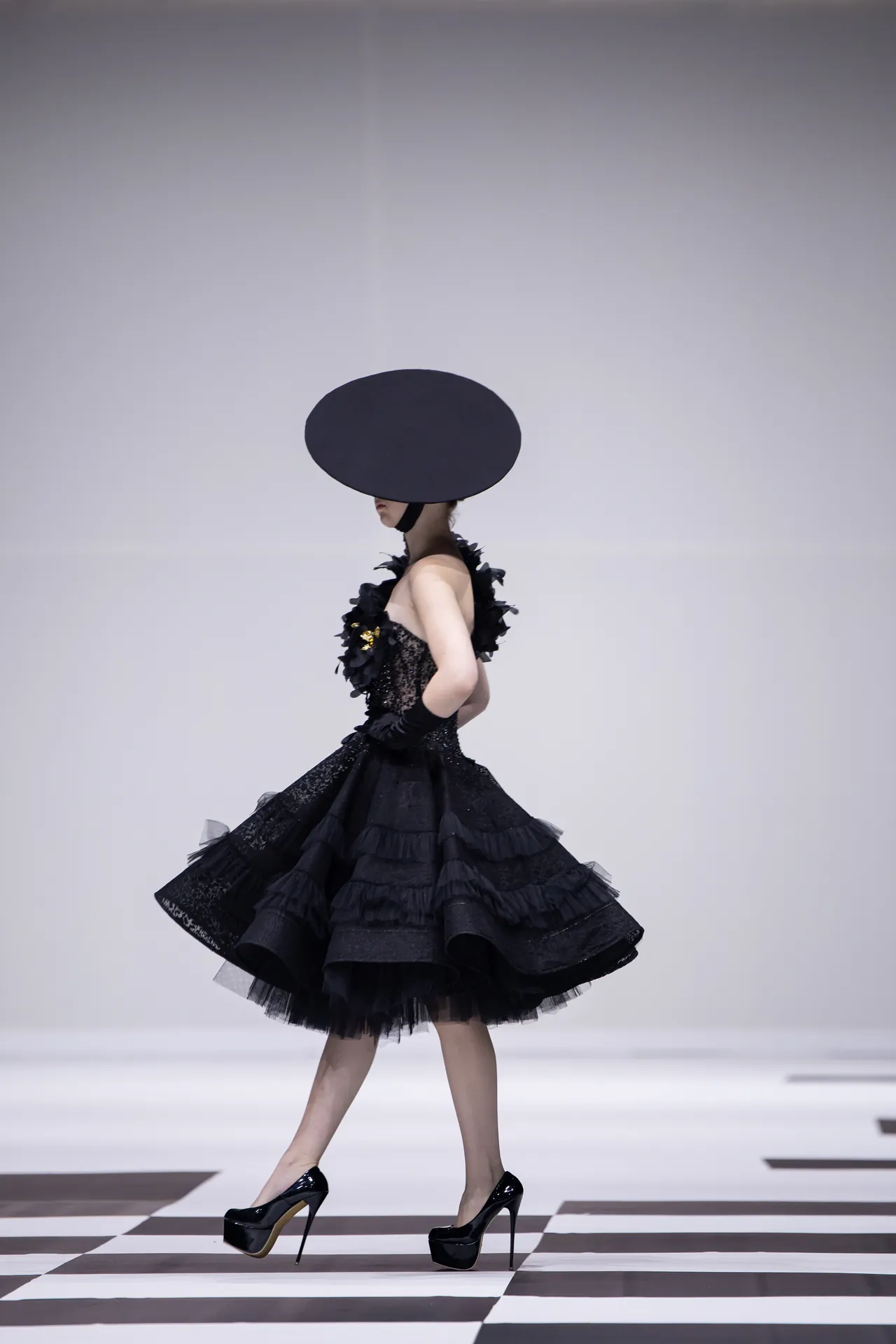 Model in a dramatic black couture gown with a large circular hat walking a checkered runway.