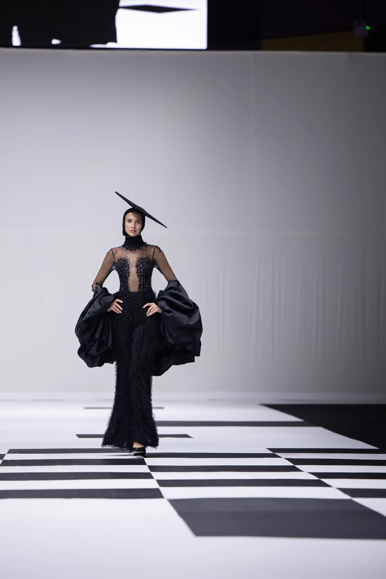 Model walking a black, floor-length gown with embellished sheer bodice and dramatic cape sleeves on a checkerboard runway.