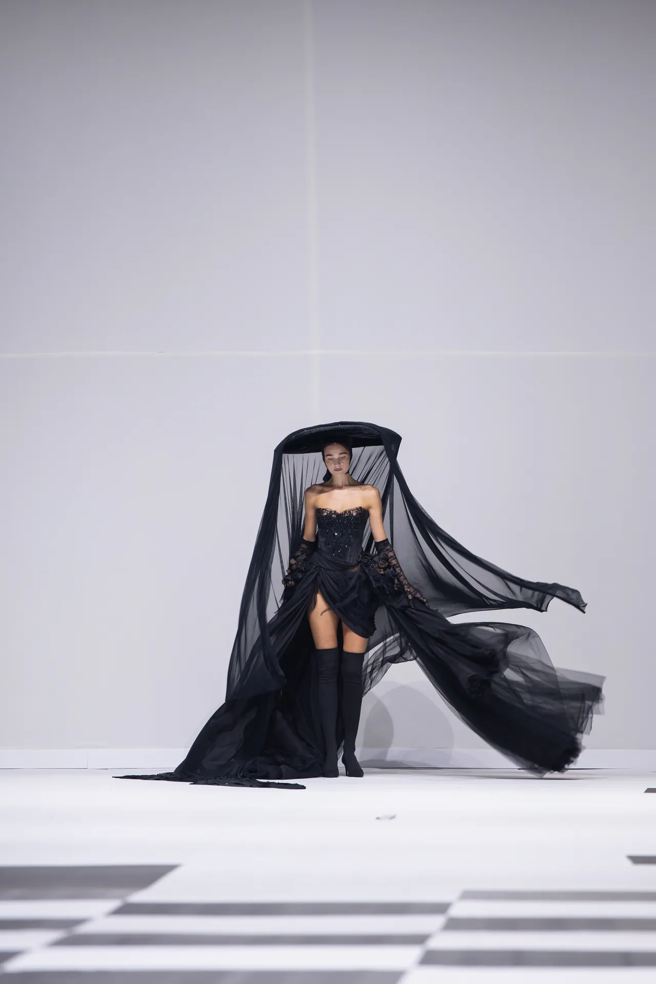 Model on a fashion runway wearing a black corset dress with a dramatic sheer cape billowing behind her against a white backdrop.
