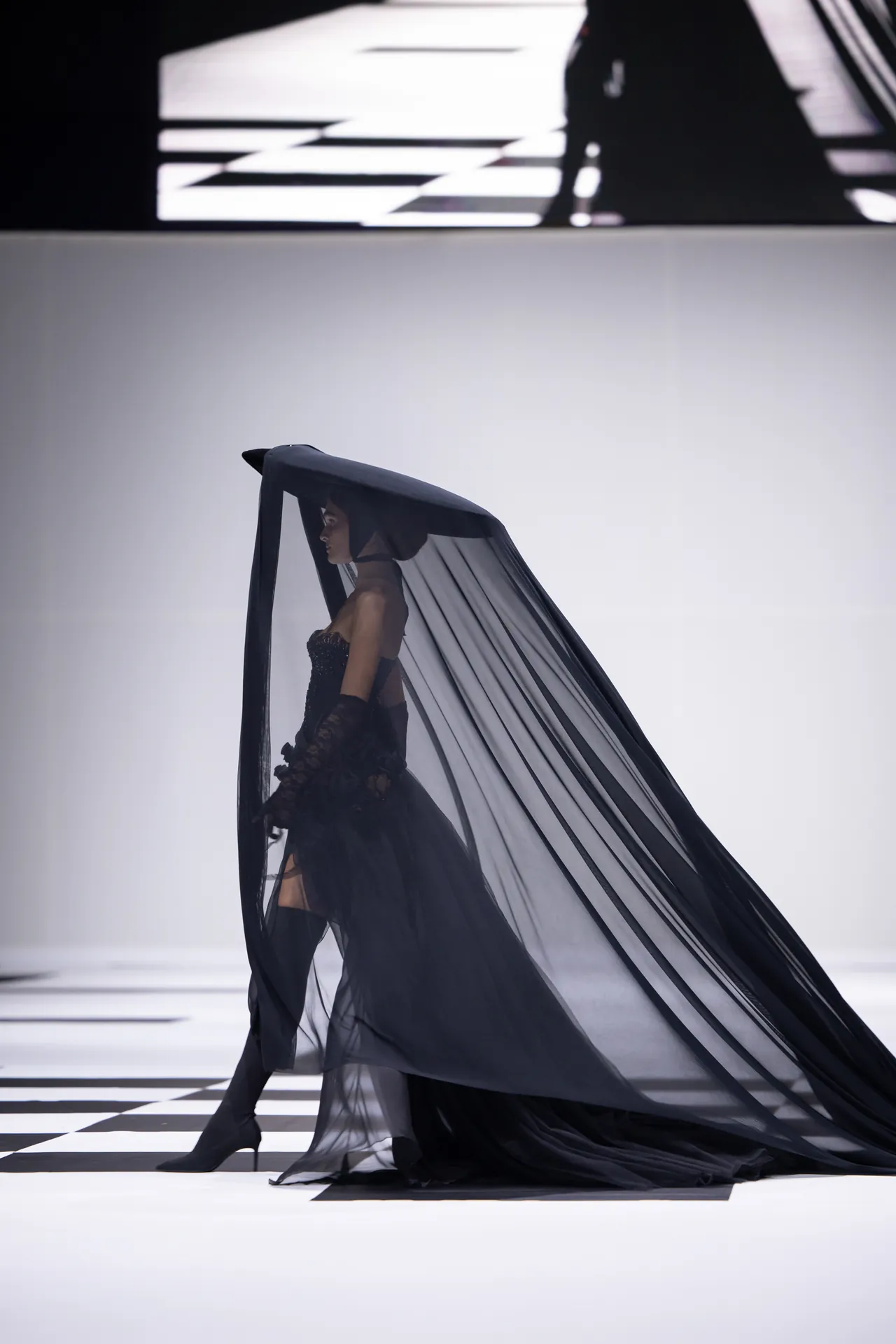 Model walking the runway in a black corseted gown with a long sheer black veil flowing behind her on a striped catwalk.
