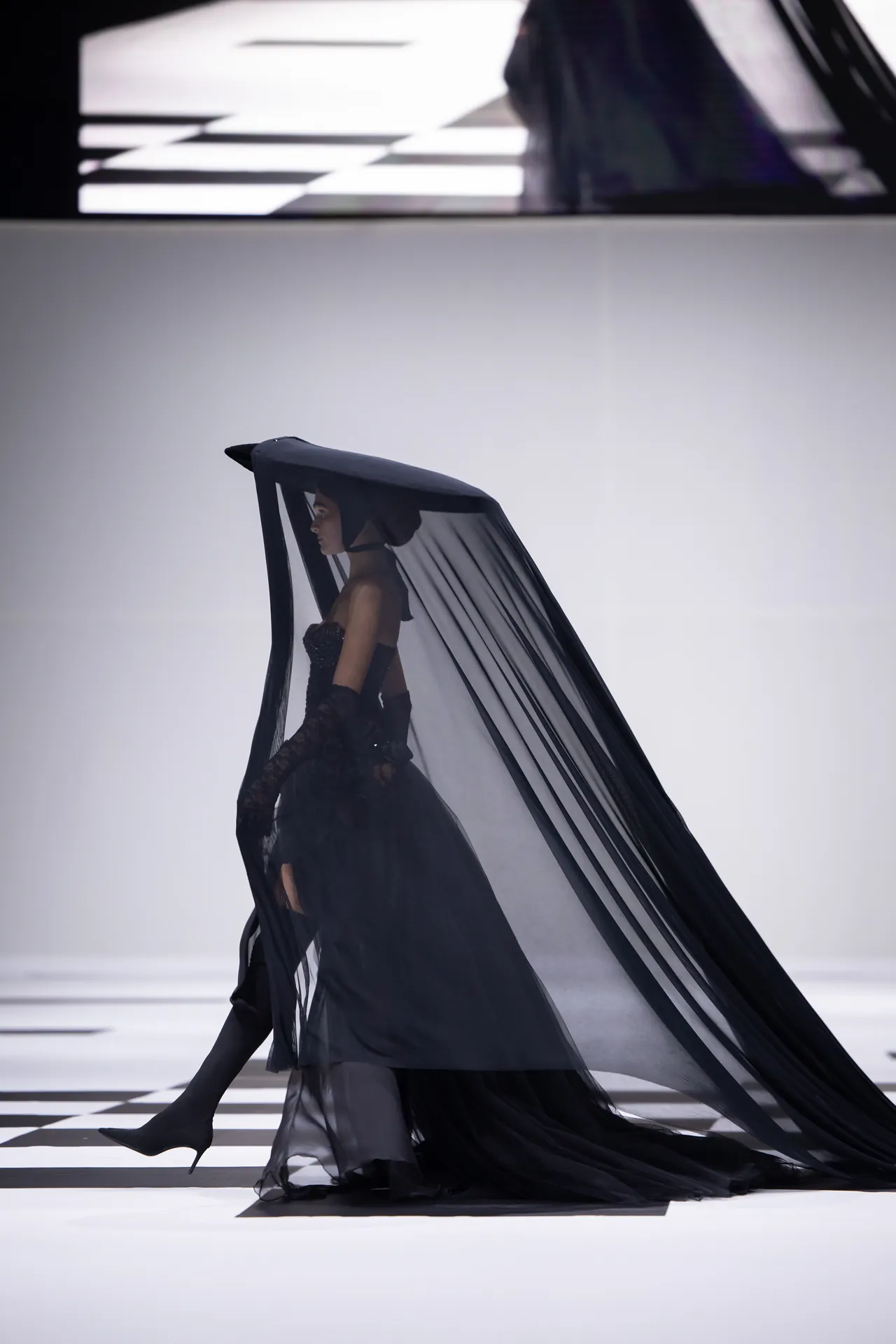 Model walking a fashion runway in a black corseted gown with a long sheer veil trailing behind her, creating a dramatic silhouette on a checkered floor.