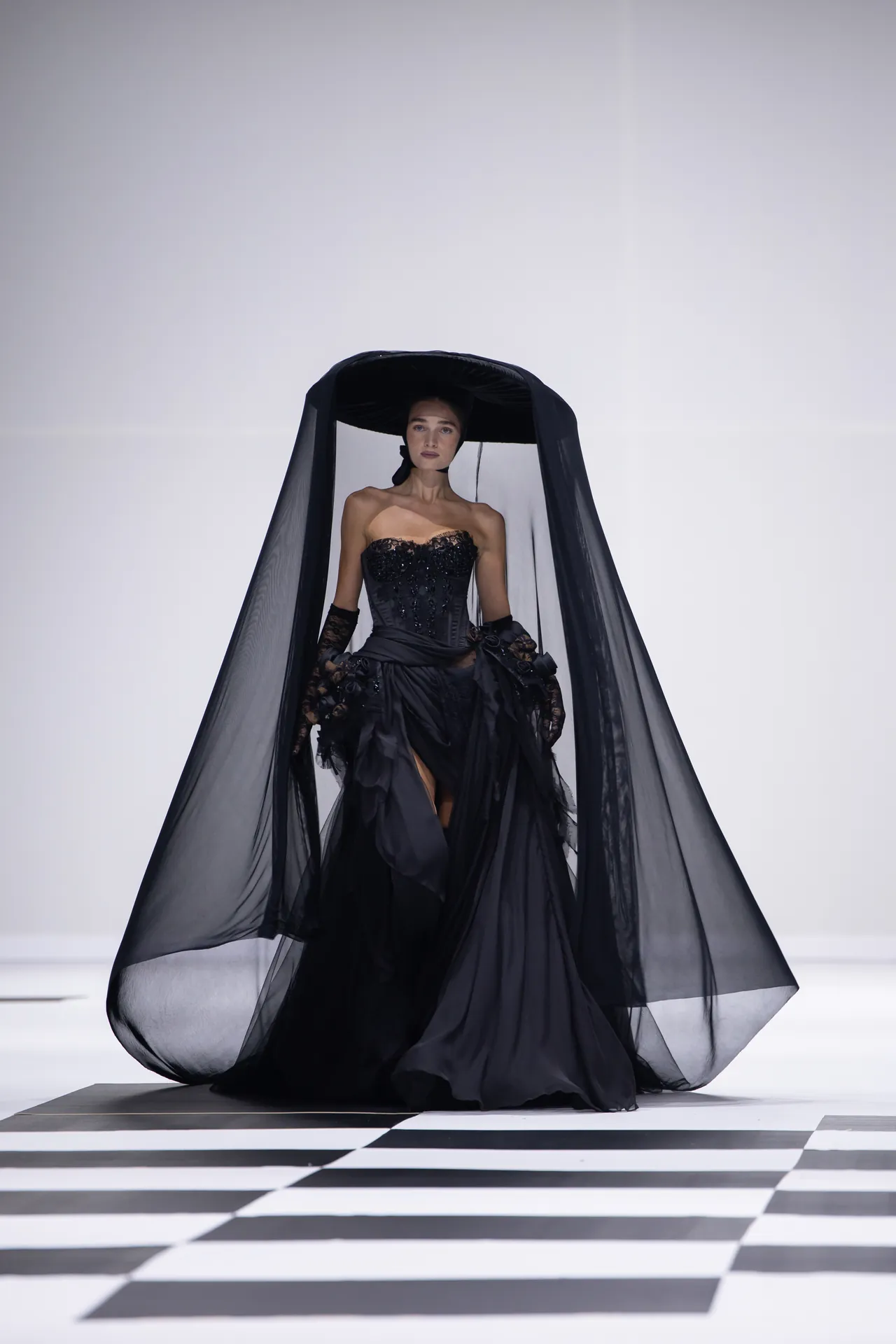 Model in a strapless black lace corset gown with a long sheer cape on a runway.