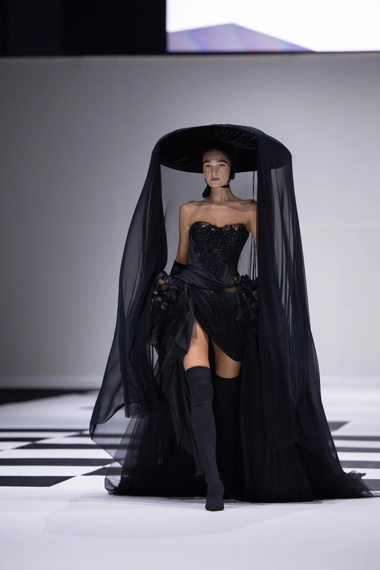 Model on a runway wearing an all-black haute-couture gown with a wide-brimmed hat and sheer veil.