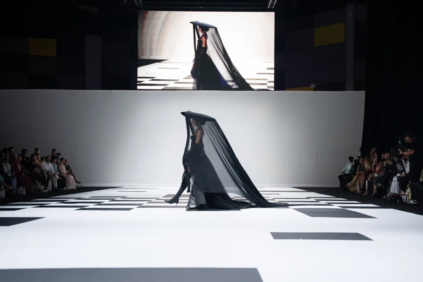 Model walks a black gown with a long sheer veil trailing behind on a geometric white runway surrounded by an audience.