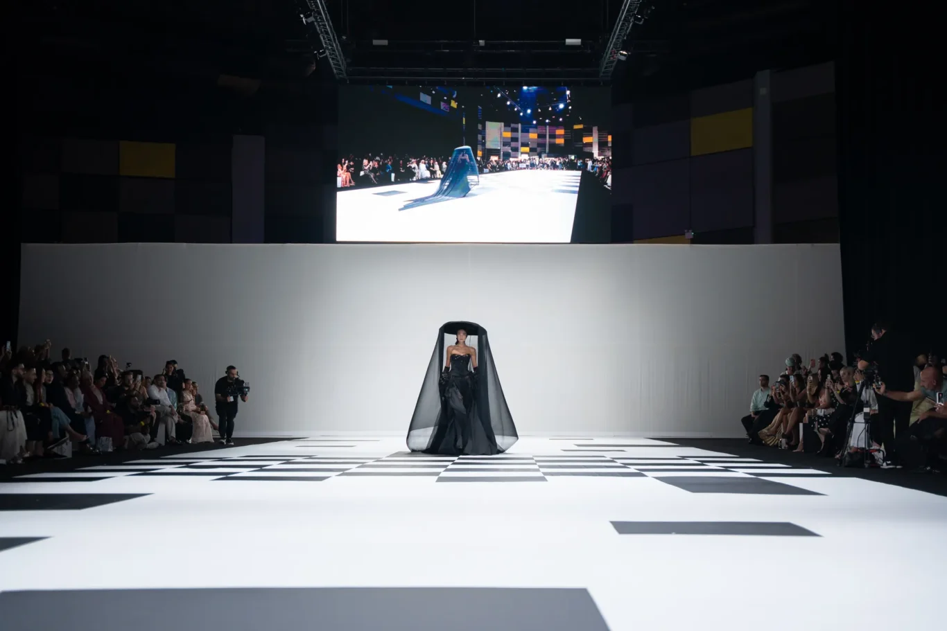 Model in a black gown with a sheer cape walks a high-fashion runway with a black-and-white checkered floor and spectators on both sides.