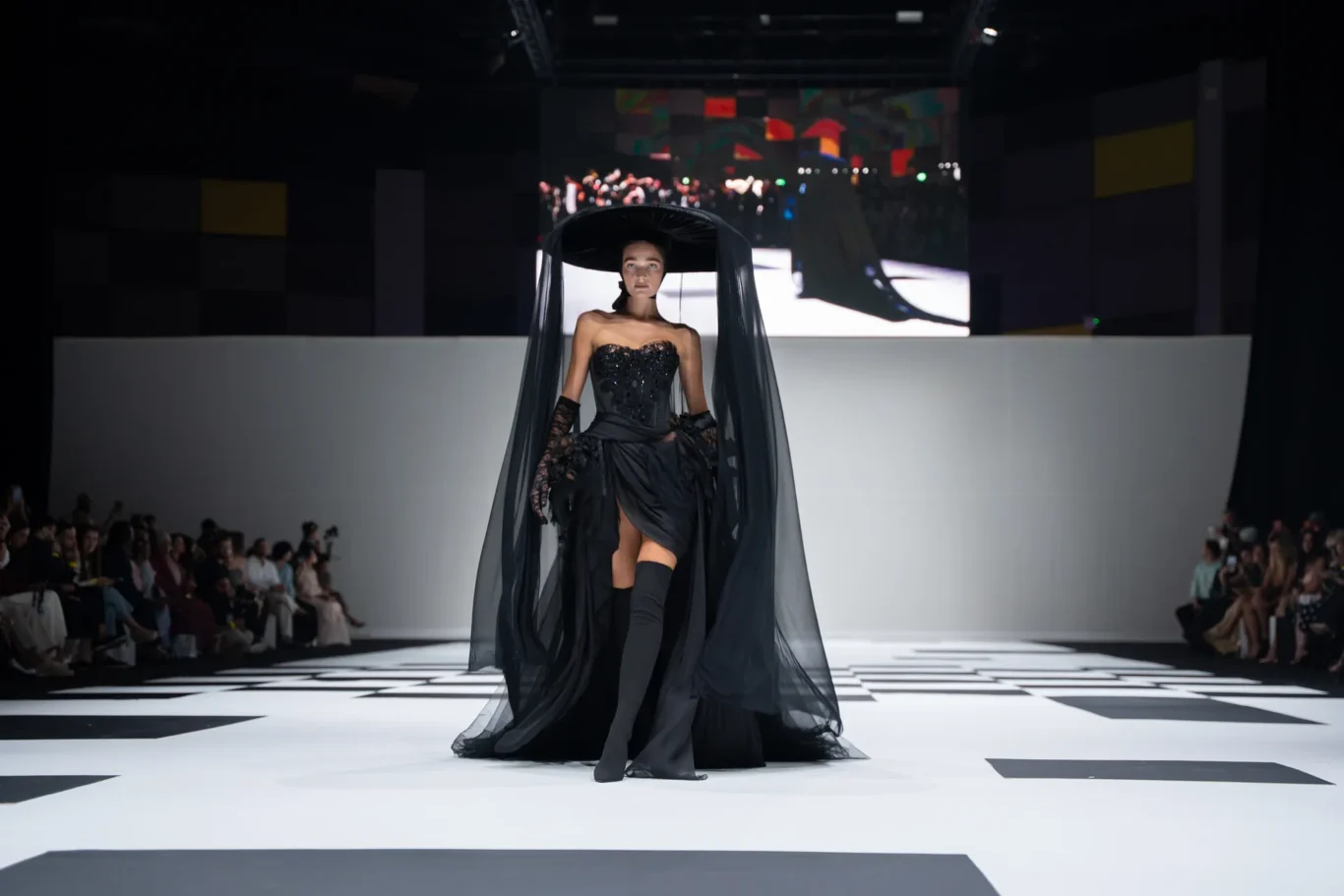 Model in a black, gothic gown with a wide veil walking a white checkered runway; audience seated along the sides.
