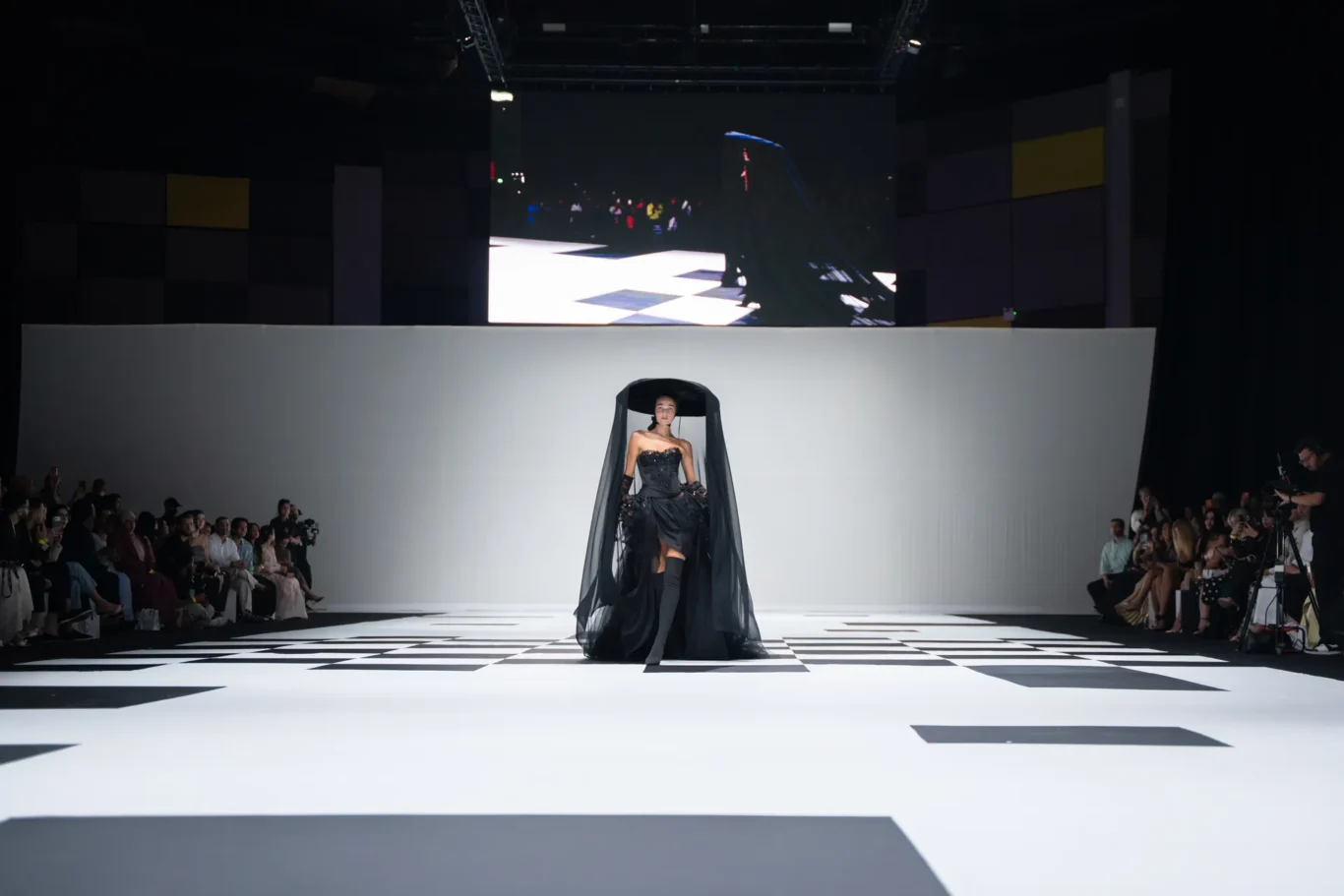 Model in an elaborate black couture dress with a sheer cape walking a black-and-white checkerboard runway; audience seated on both sides.