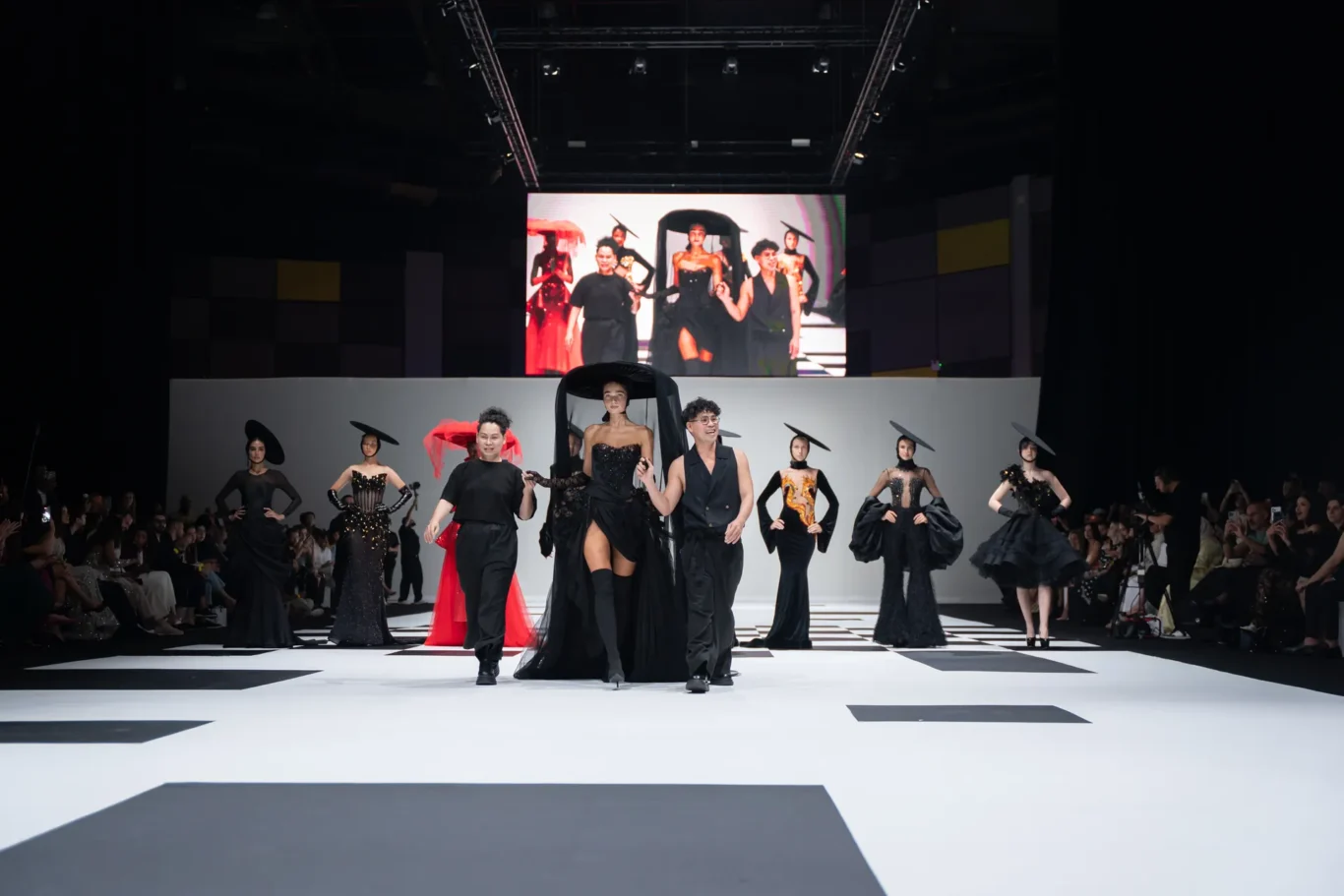 Models walk a couture runway in black gowns with red accents as a large screen shows the show in the background.