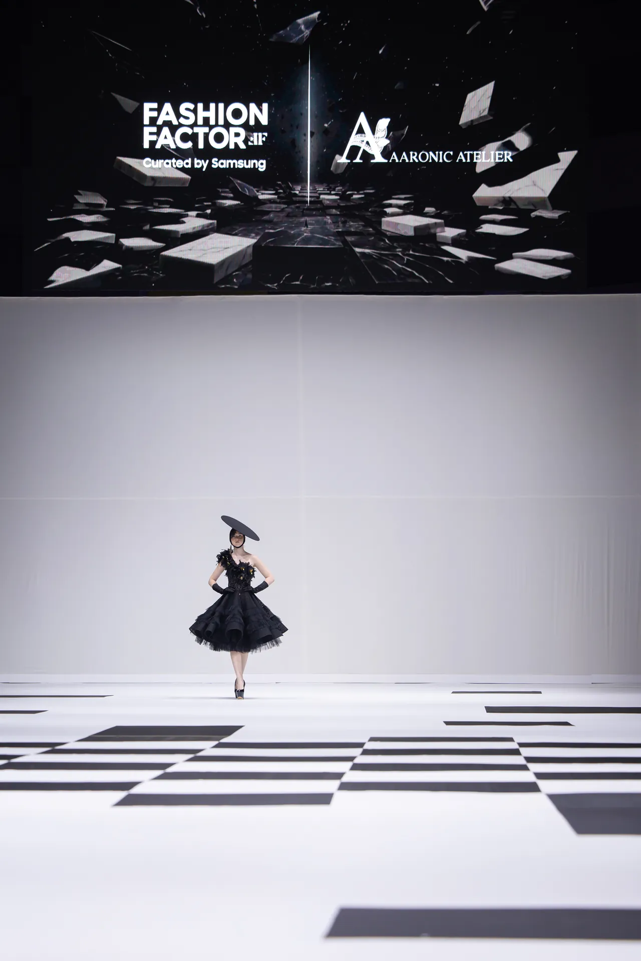 Model walking the runway in a black voluminous gown with a large circular hat on a white geometric-stage floor.