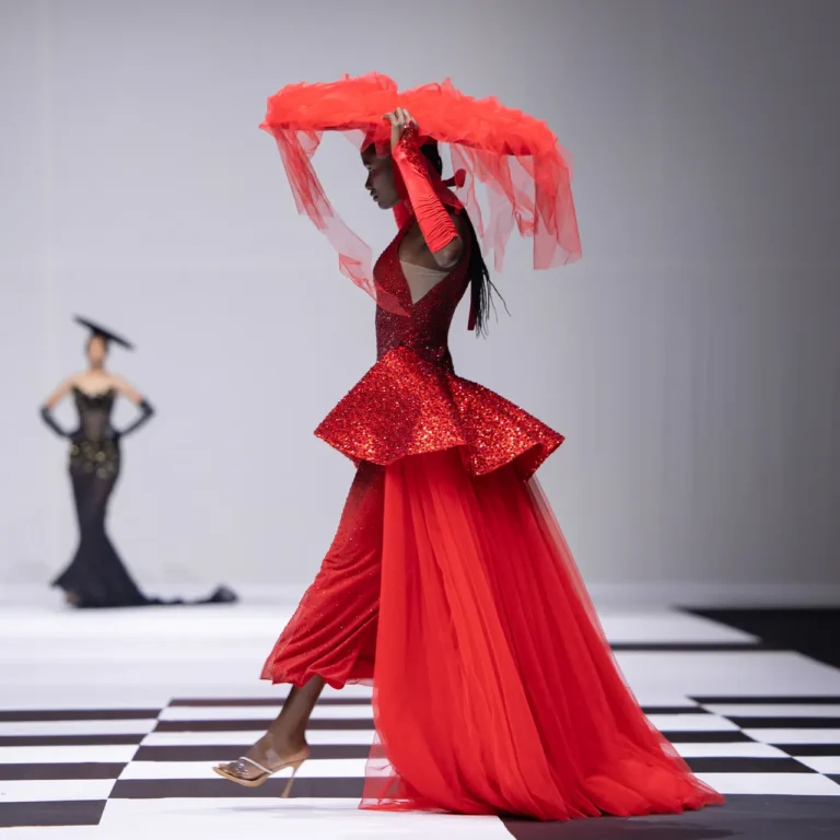 Model wearing a red glitter gown on a fashion runway, holding a red veil above her head.