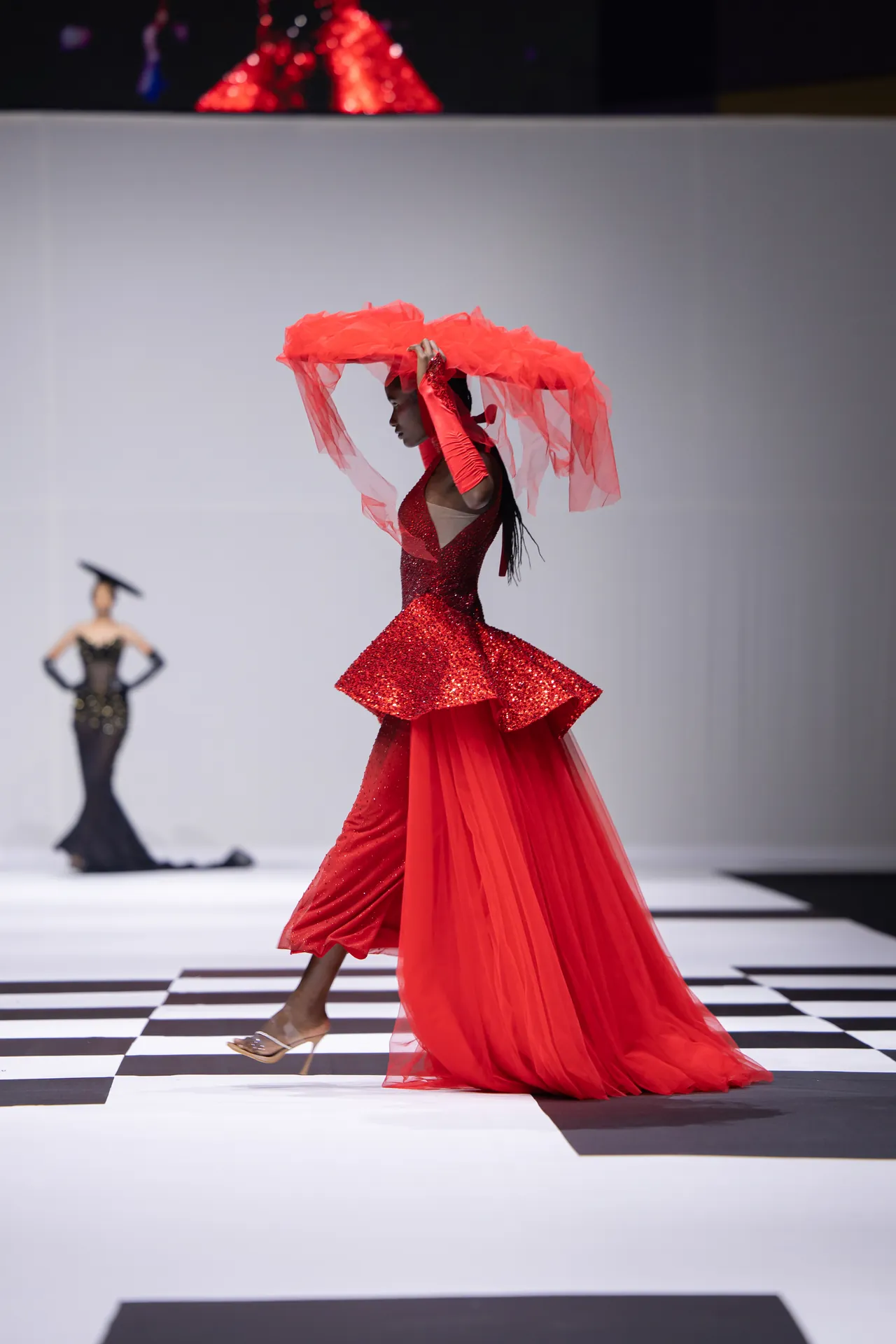 Model wearing a red glitter gown on a fashion runway, holding a red veil above her head.