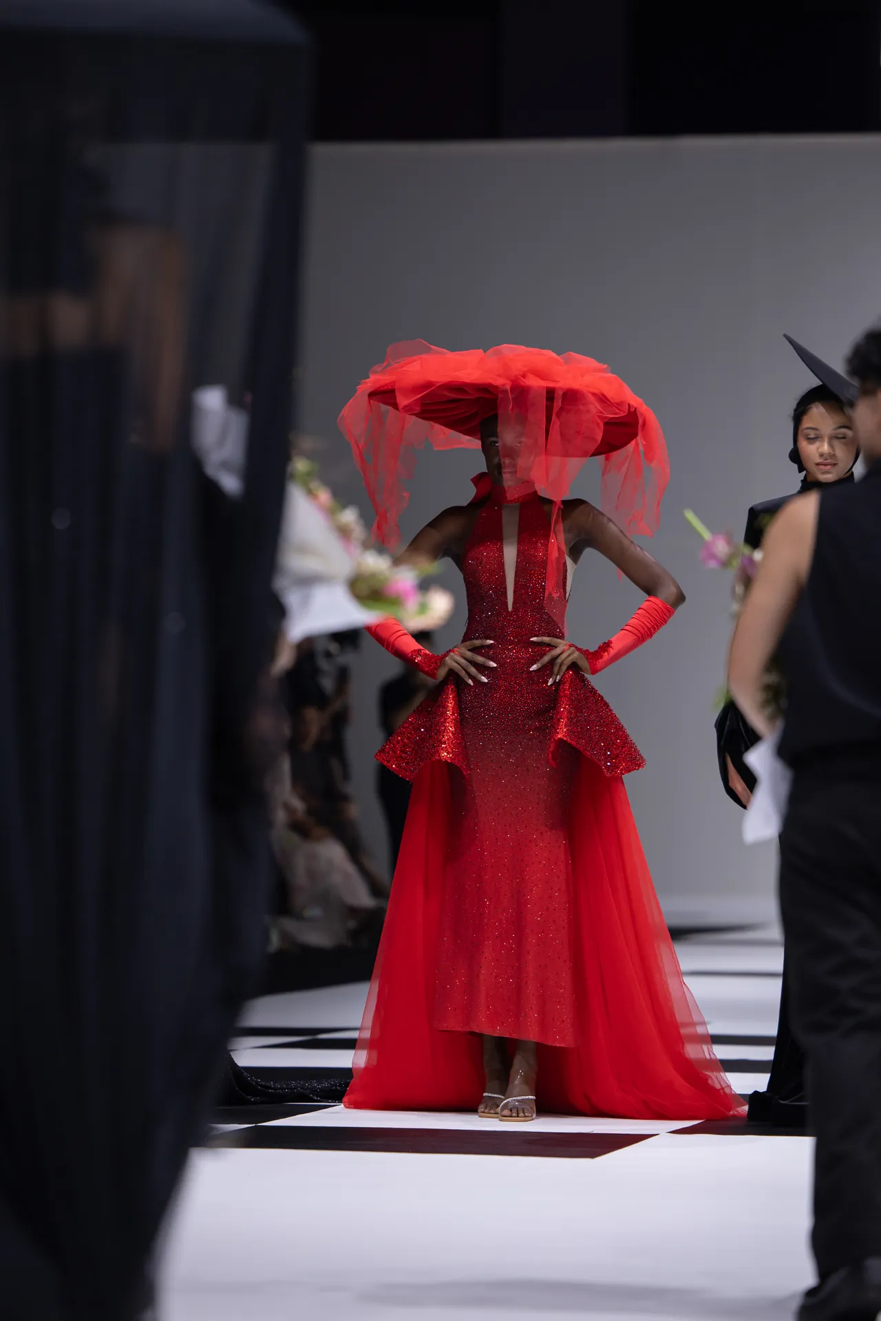Model on a runway wearing a sparkling red gown with a large red feathered hat; hands on hips.