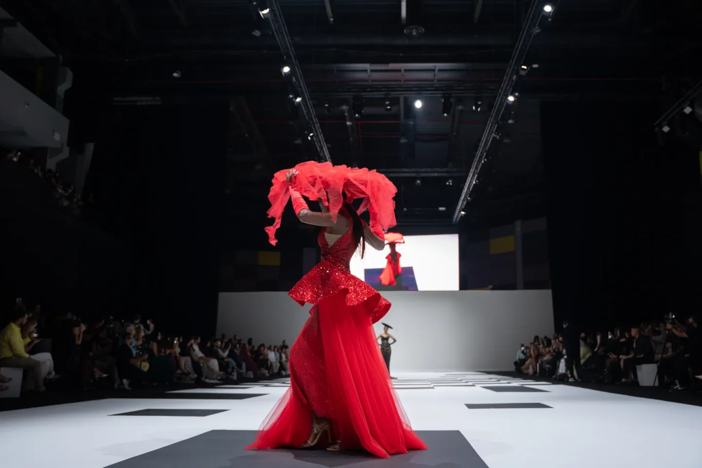 Model wearing a red sequined gown on a runway, lifting billowing red fabric above her head as spectators watch.