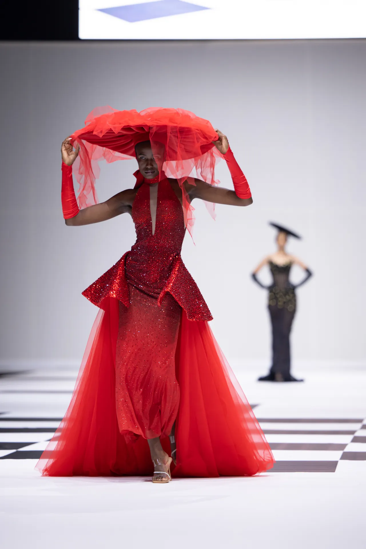 Model in a sparkling red gown on a runway, lifting a voluminous red tulle headpiece above her head.