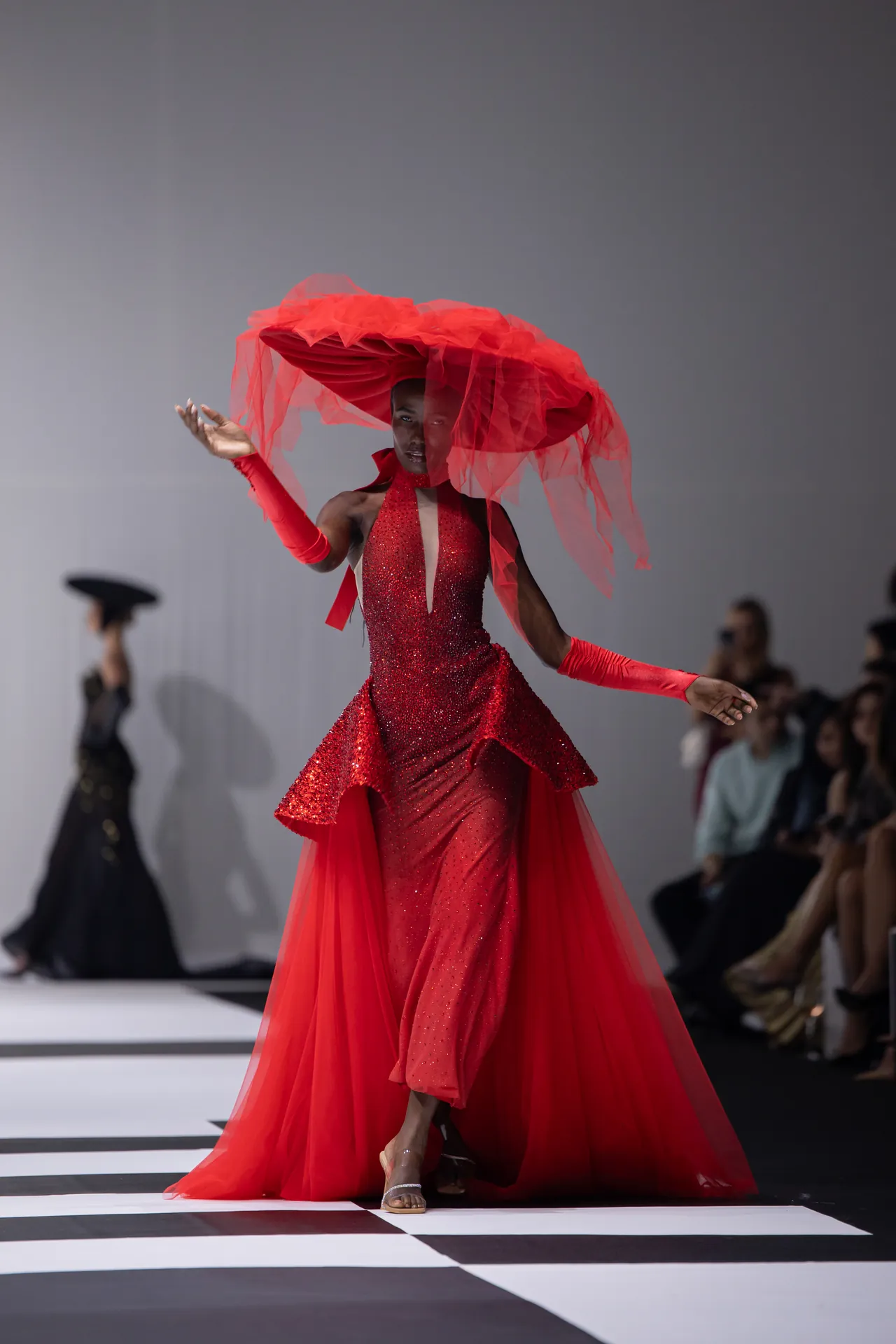 Model in a red sequined gown with a voluminous red veil headpiece walking a striped runway on a fashion show stage.