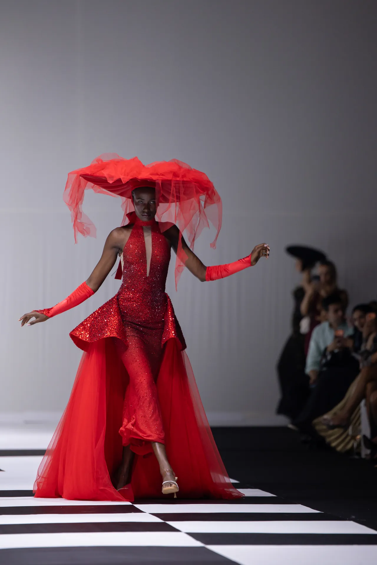 Model walks the runway in a red sequined gown with a voluminous red tulle headpiece and flowing cape, audience in the background.