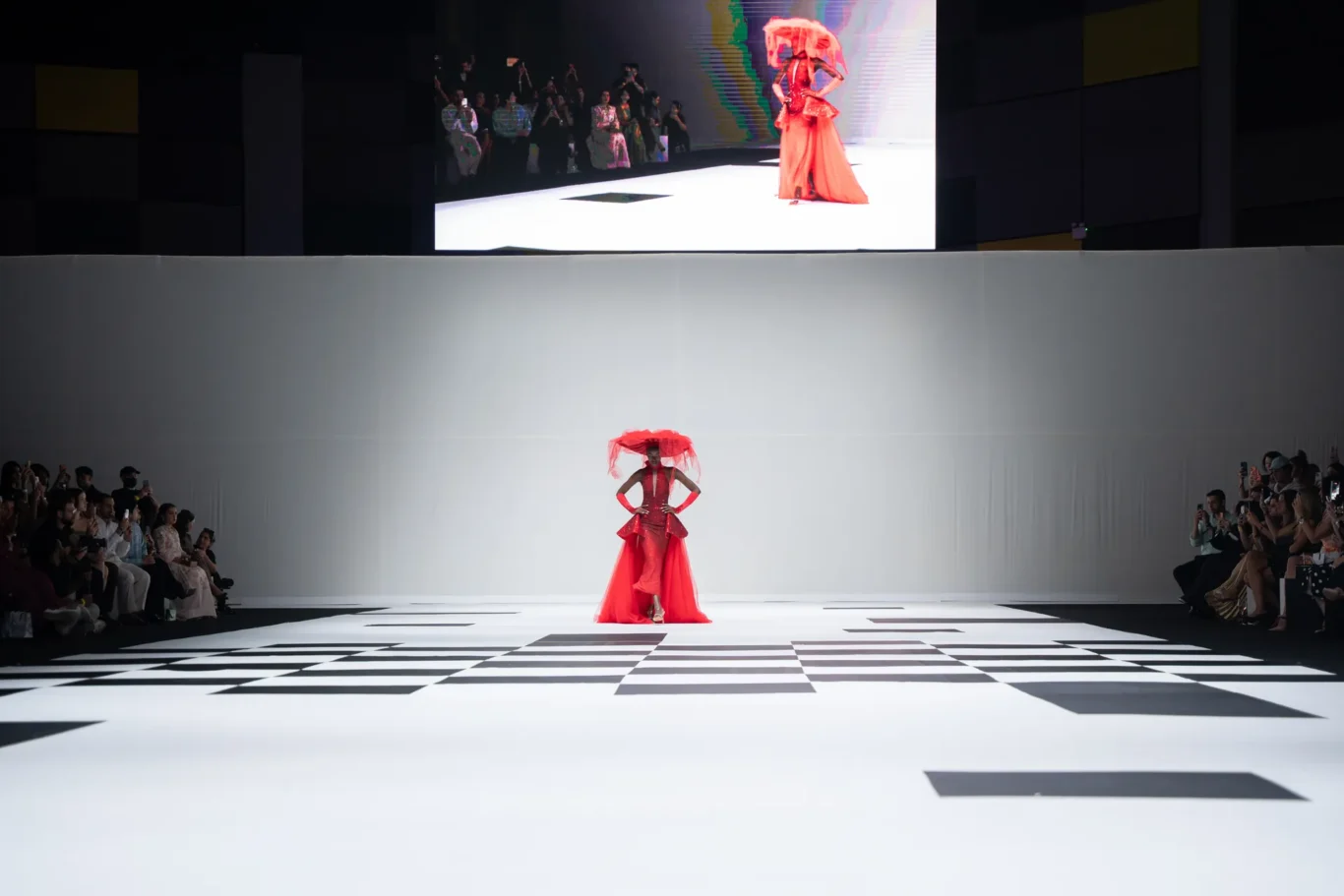 Model in a red gown with a sheer red veil walks a black-and-white checkered runway, audience on both sides watching.