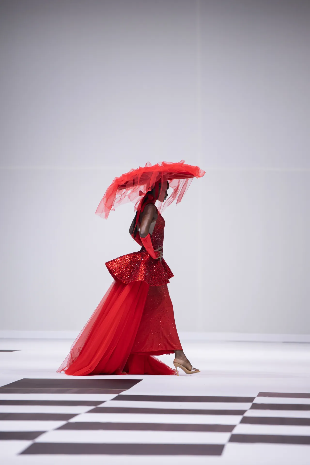 Model walks a runway in a floor-length red sequined gown with a voluminous red tulle headpiece and flowing train.