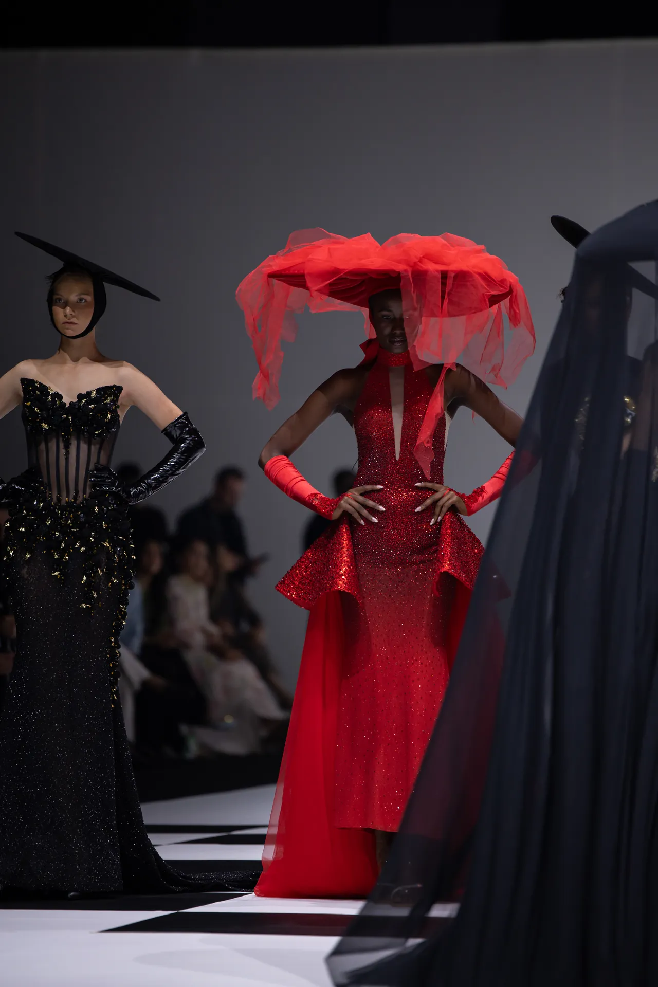 Models walk a fashion runway in dramatic gowns; center model wears a red sparkling gown with a huge red veil-like headpiece, flanked by black-glittering dress on left.