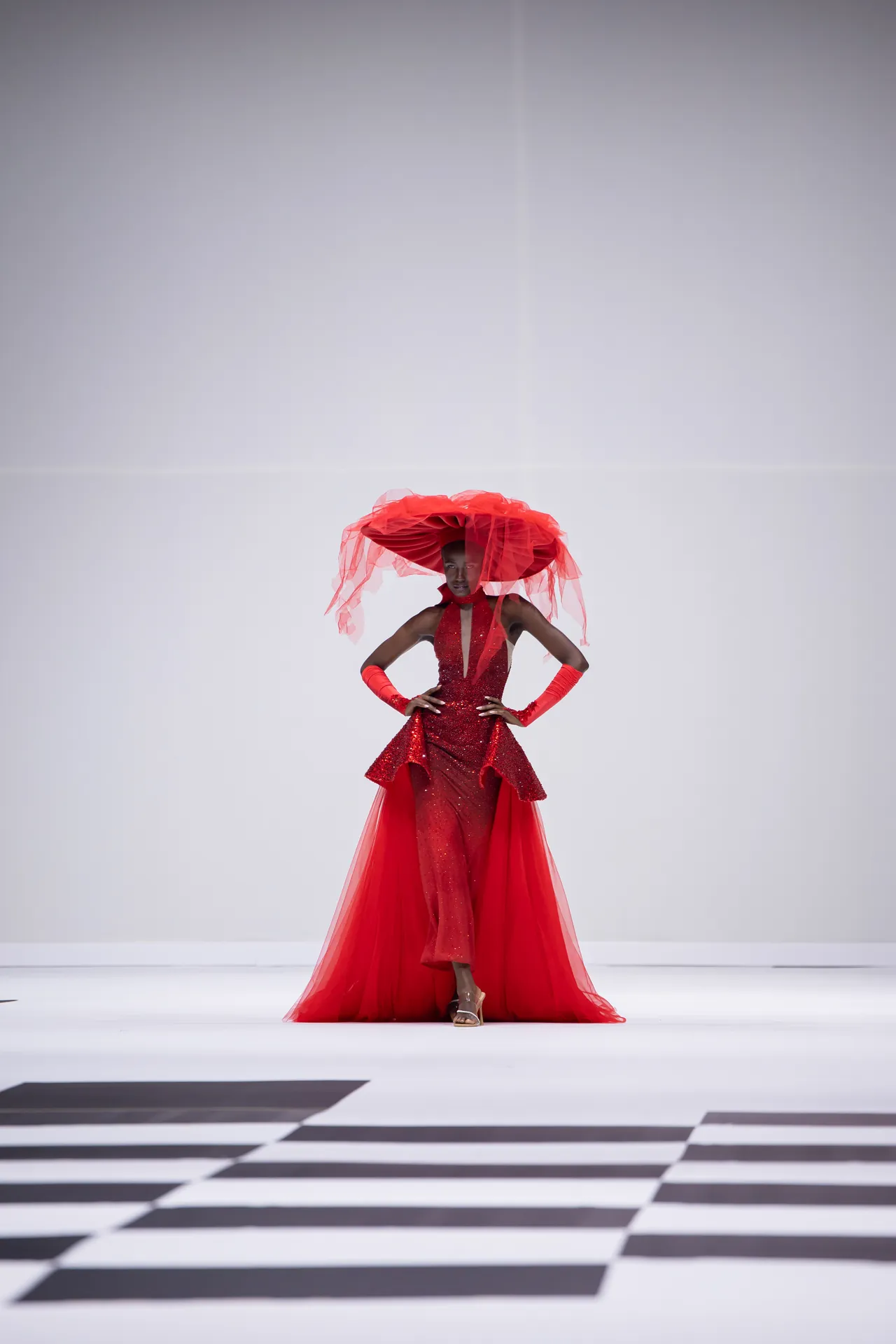 Model in a glittering red gown with a dramatic red headpiece on a fashion runway.