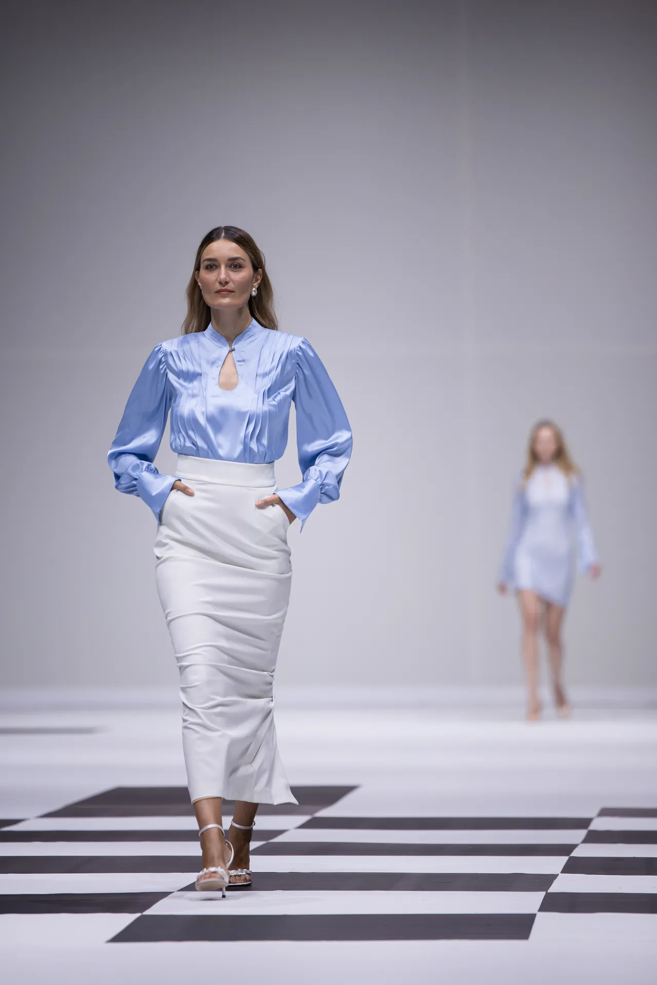 Model walking a fashion show on a black-and-white checkered runway, wearing a light blue blouse and white pencil skirt.