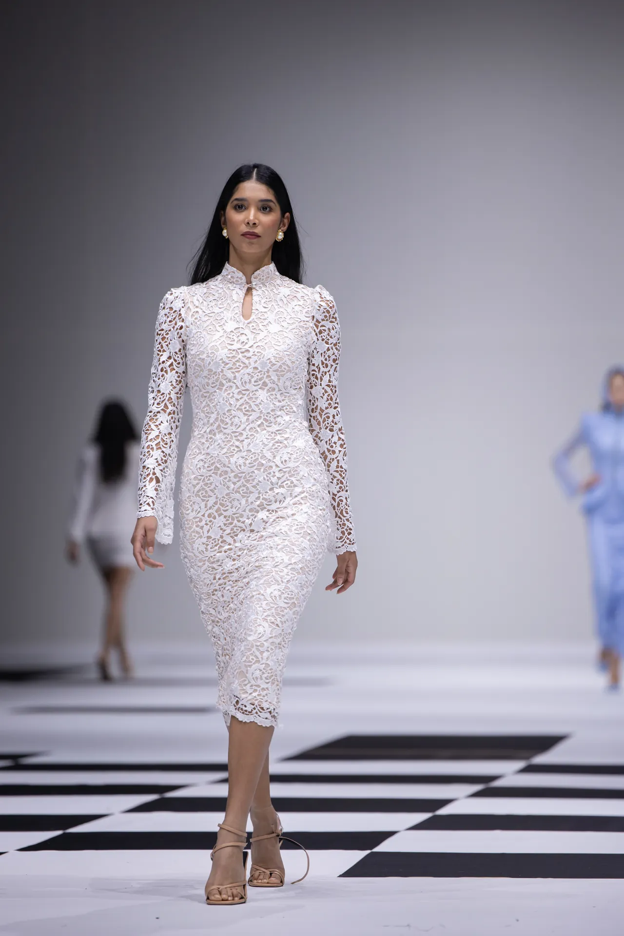 Model walking a fashion runway in a knee-length white lace dress with floral pattern and beige heels.