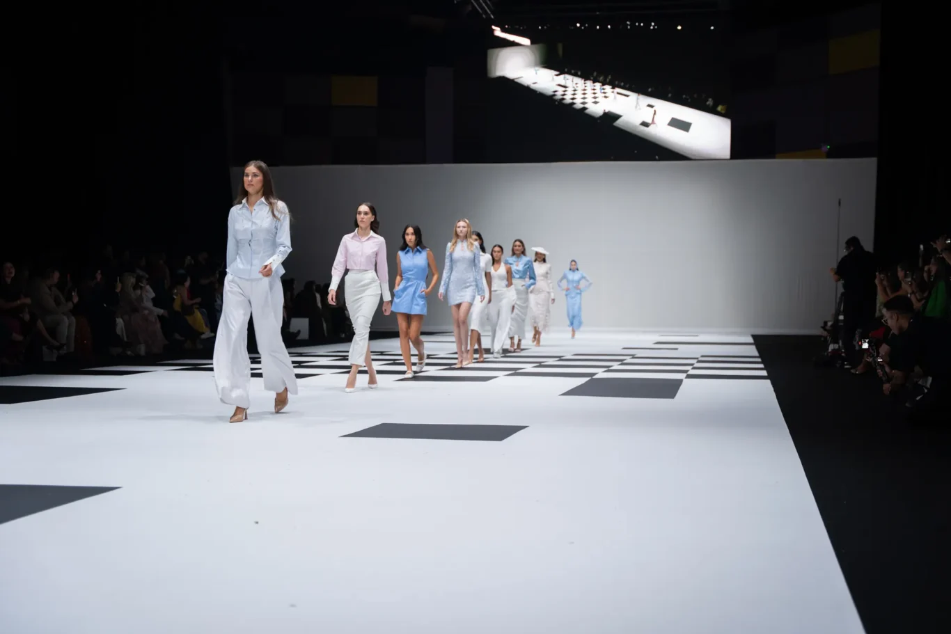 Models stride down a white runway with black geometric tiles at a fashion show, wearing pastel blue and white outfits.