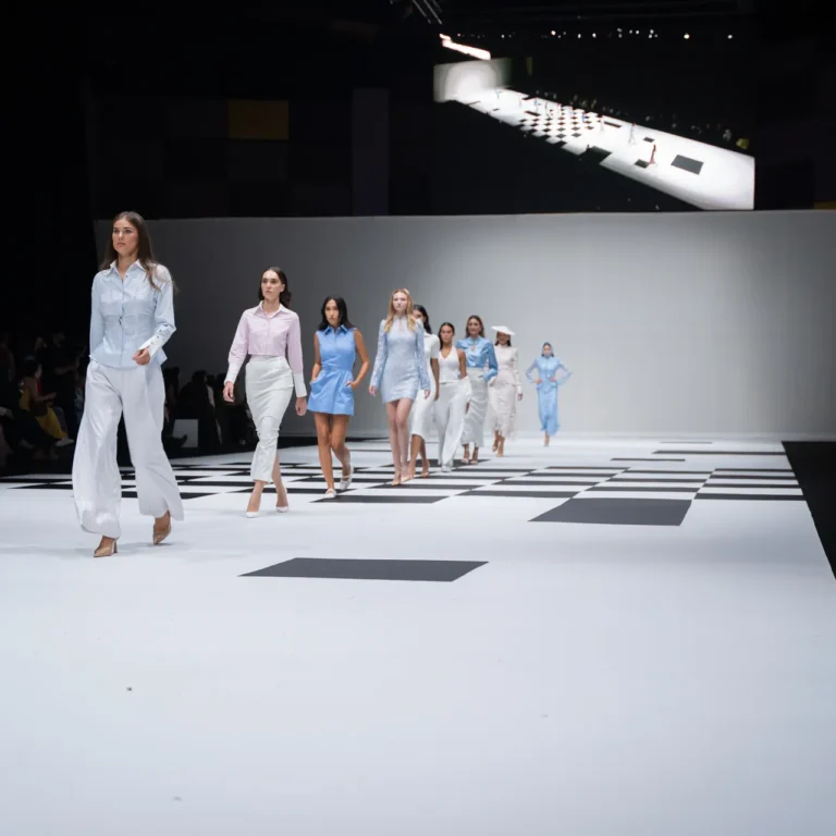 Models stride down a white runway with black geometric tiles at a fashion show, wearing pastel blue and white outfits.