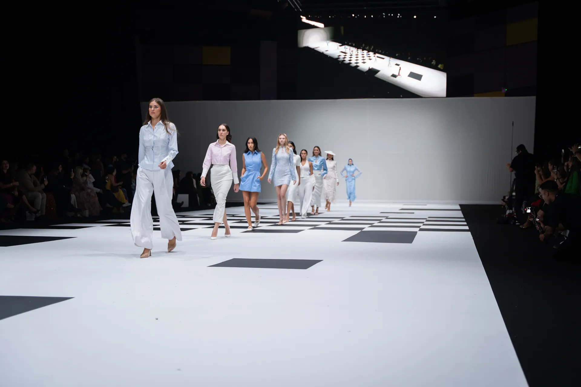 Models stride down a white runway with black geometric tiles at a fashion show, wearing pastel blue and white outfits.