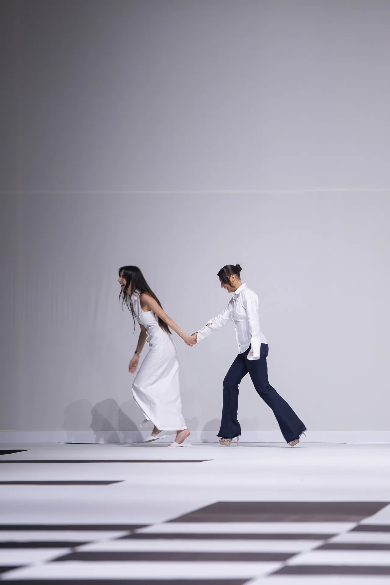 Two models walk hand-in-hand on a minimalist white runway with a black-and-white checkered floor.