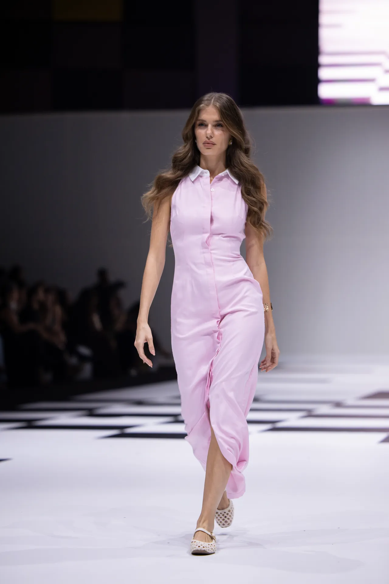 Female model wearing a light pink sleeveless button-down jumpsuit walking down a fashion runway with a checkered floor pattern behind her in white lights.