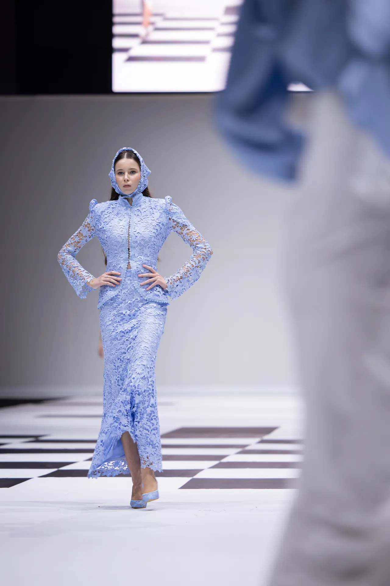 Model wearing a light blue lace hooded dress walking a black-and-white checkered runway.