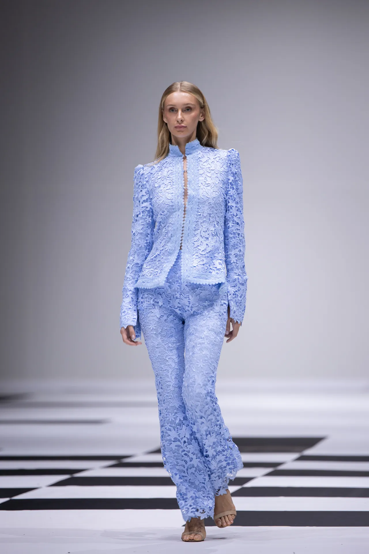 Runway model walking in a pale blue lace pantsuit with floral pattern.
